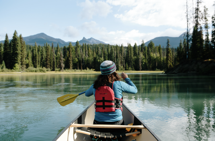 A woman in a canoe paddling on a calm river surrounded by a forested landscape with mountains in the background.