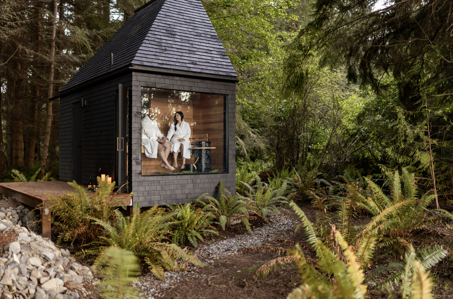 Two people sitting inside a small black wooden sauna with glass window, surrounded by lush green forest.