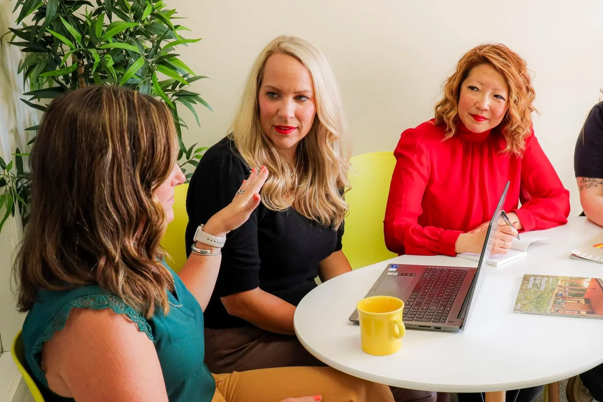 Women having a discussion around a table with a laptop, books, and a yellow mug in a meeting room with green plants.