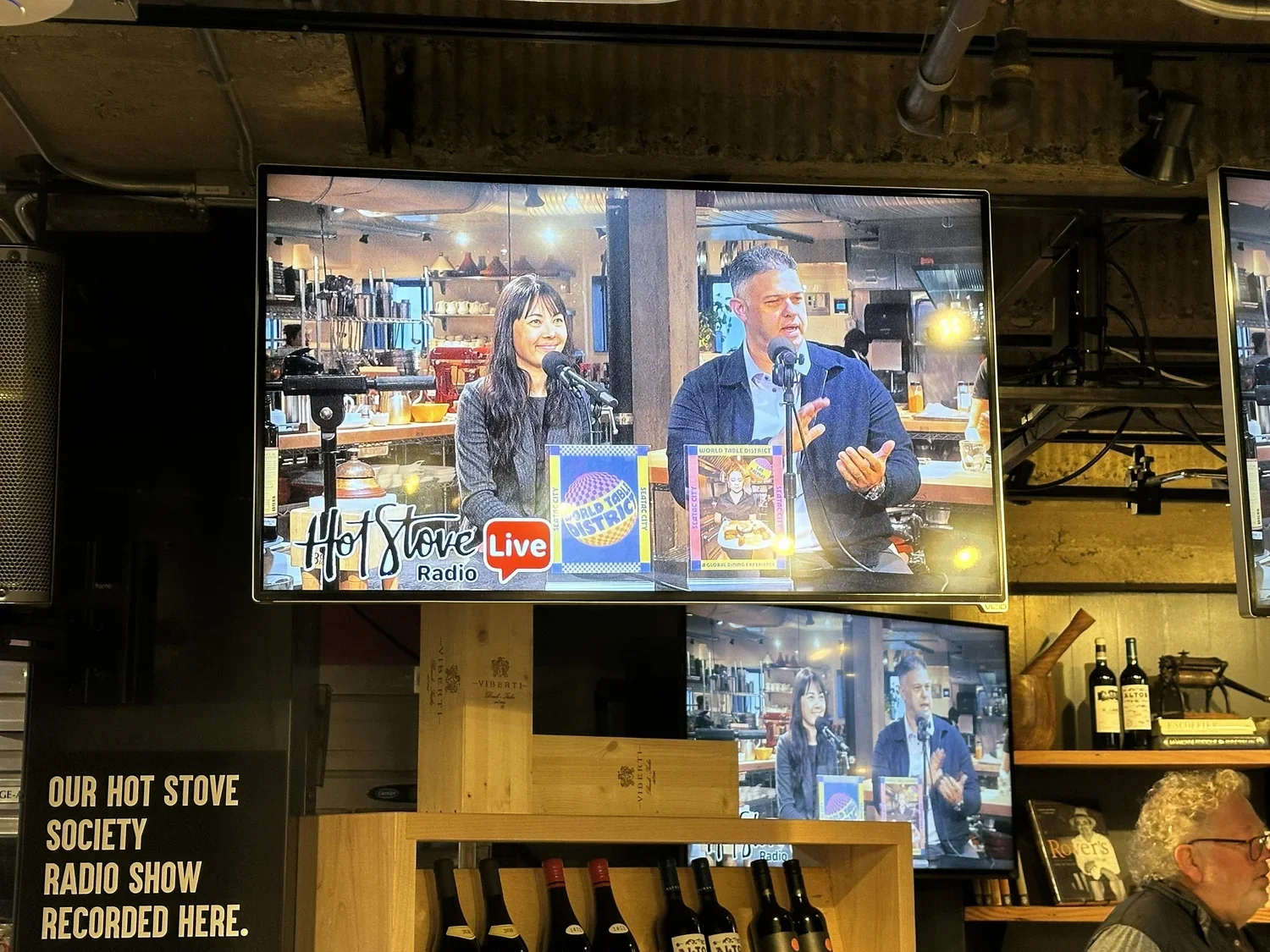 Two people speaking into microphones on a stage or in a studio, with shelves of kitchen items behind them. The screen displays a live broadcast for Hot Stove Radio with the logo and the words "Live".
