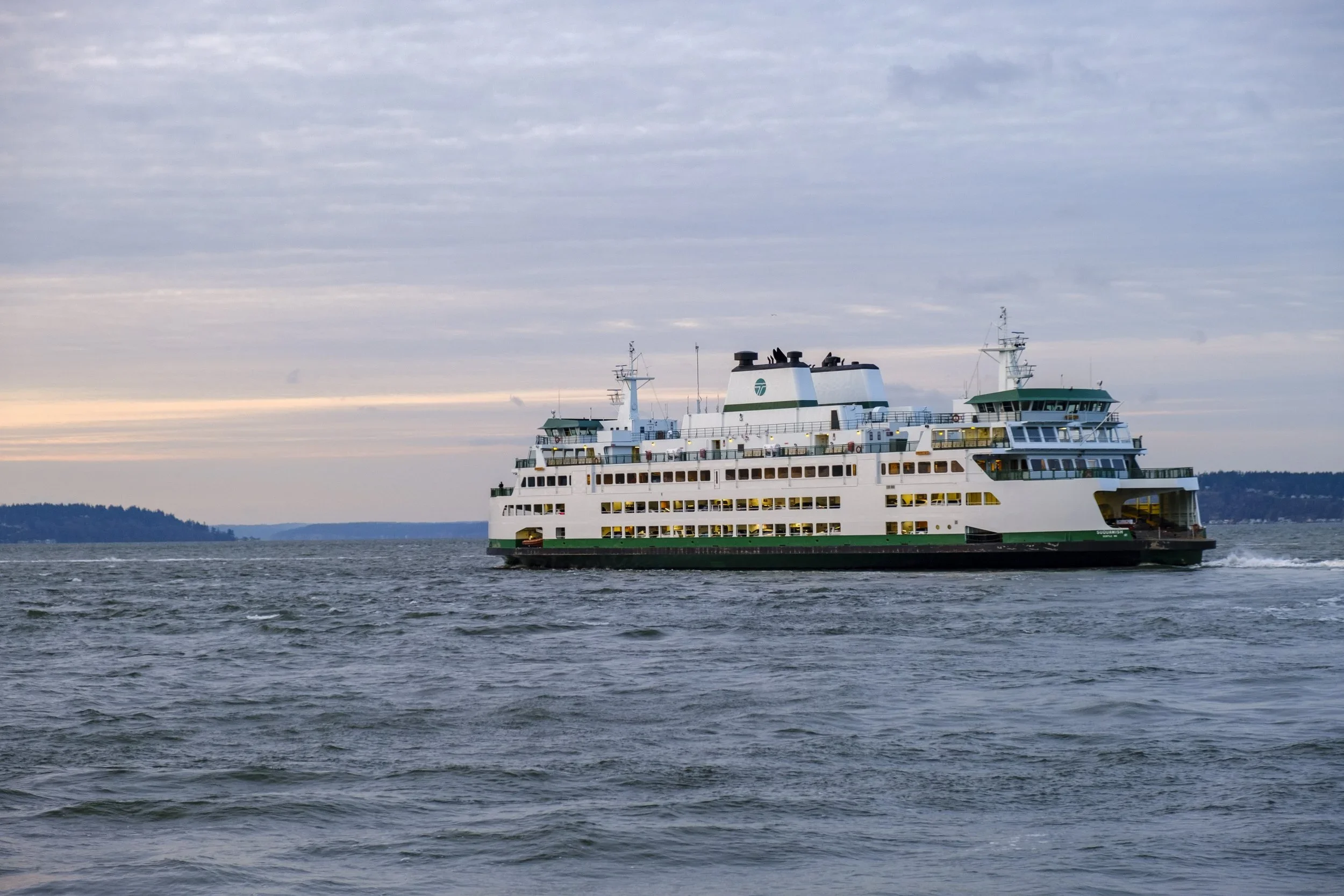 A large passenger ferry boat sailing on the water under a cloudy sky with distant land in the background.