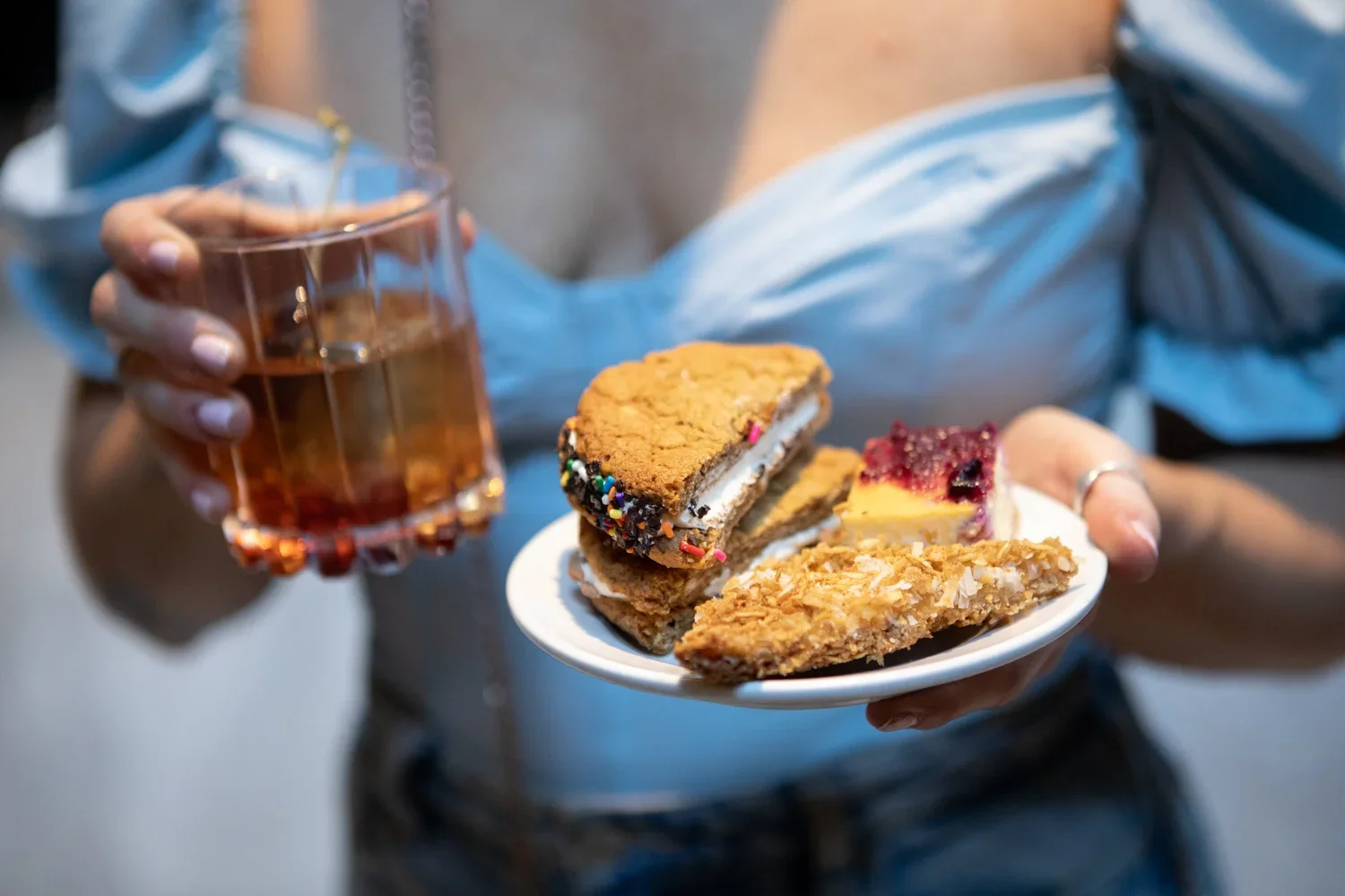 A person holding a glass of rose-colored drink in one hand and a small white plate with assorted desserts in the other. The desserts include an ice cream sandwich, a slice of berry-topped cheesecake, and a flat pastry.