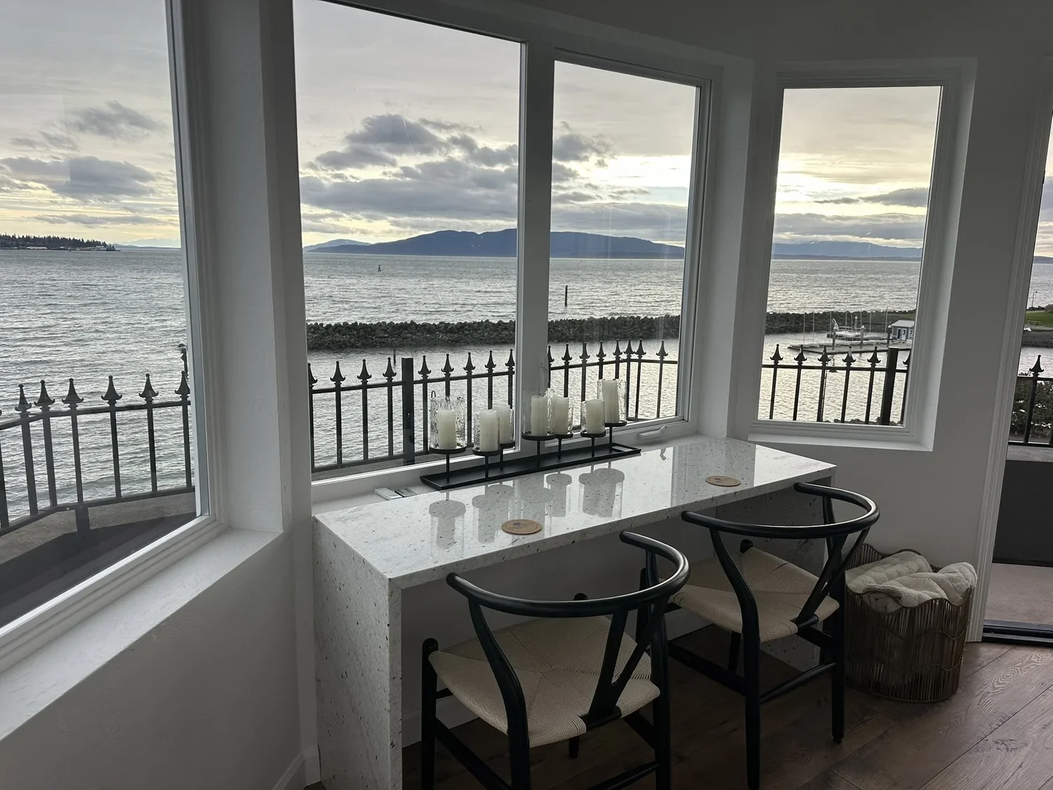 Interior view of a room with large windows overlooking a body of water, mountains in the distance, and cloudy sky; a white speckled stone window seat with candles and two chairs in front.