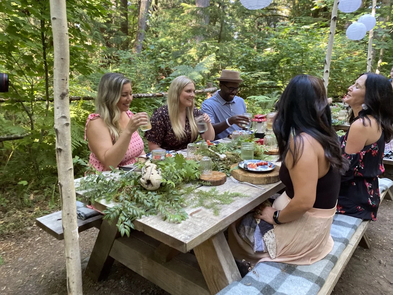 A group of six women and one man are sitting at a wooden picnic table outdoors in a lush forest, enjoying food and drinks. They are smiling and engaged in conversation, with decorations and greenery on the table.