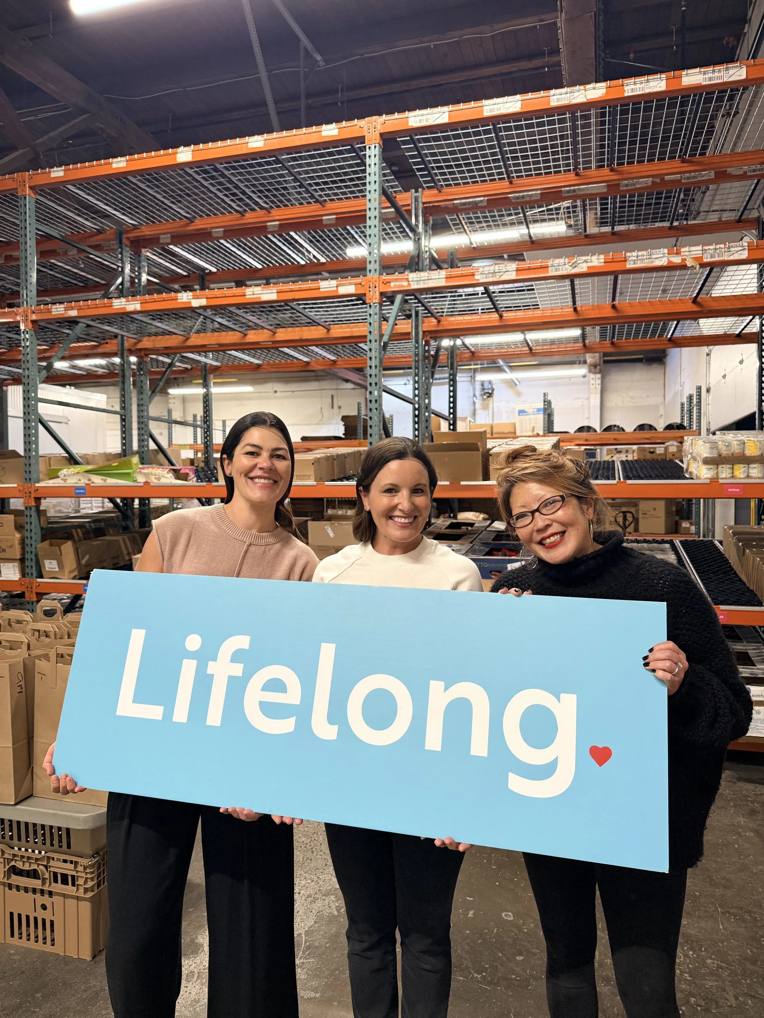 Three women smiling and holding a blue Sign with the word 'Lifelong' and a small red heart in a warehouse setting.
