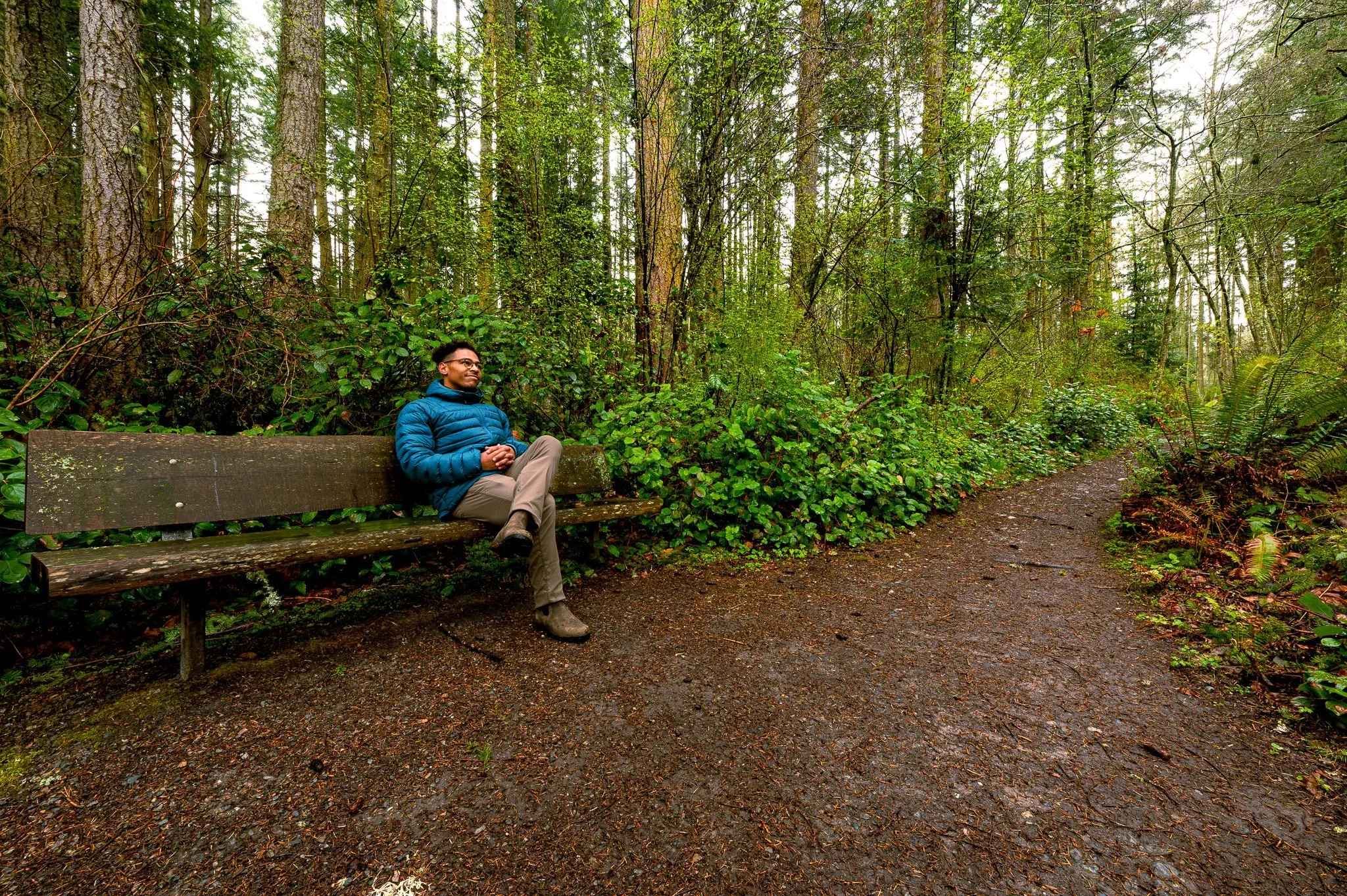 A man sitting on a wooden bench in a lush green forest with tall trees and dense foliage, wearing glasses, a blue puffer jacket, and khaki pants, smiling and relaxing.