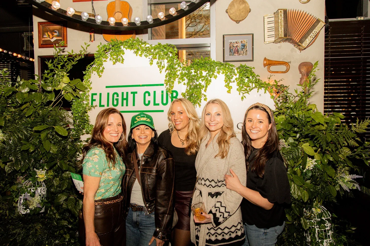 Group of five women smiling and posing in front of a lush green backdrop with a sign that reads 'Flight Club' at an indoor event space decorated with musical instruments and greenery.