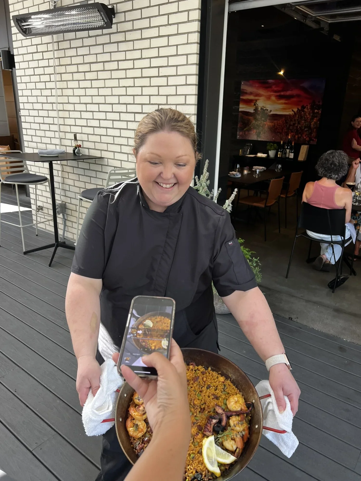 A woman dressed as a chef stands holding a large pan of seafood paella at an outdoor dining area. In the foreground, a person is taking a photo of her with a smartphone. There are tables and chairs with some people dining in the background.