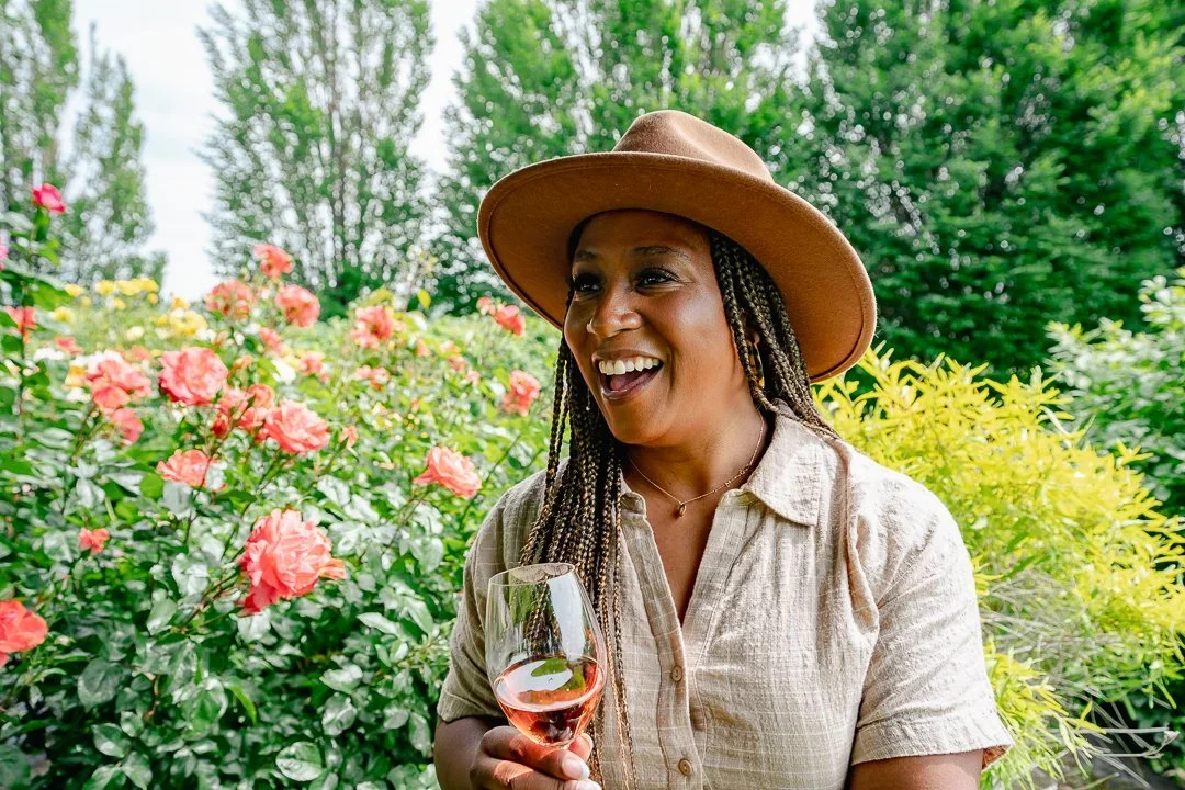 Woman with braids wearing a wide-brimmed hat and beige shirt, smiling and holding a glass of rosé wine outdoors among pink roses and green foliage.