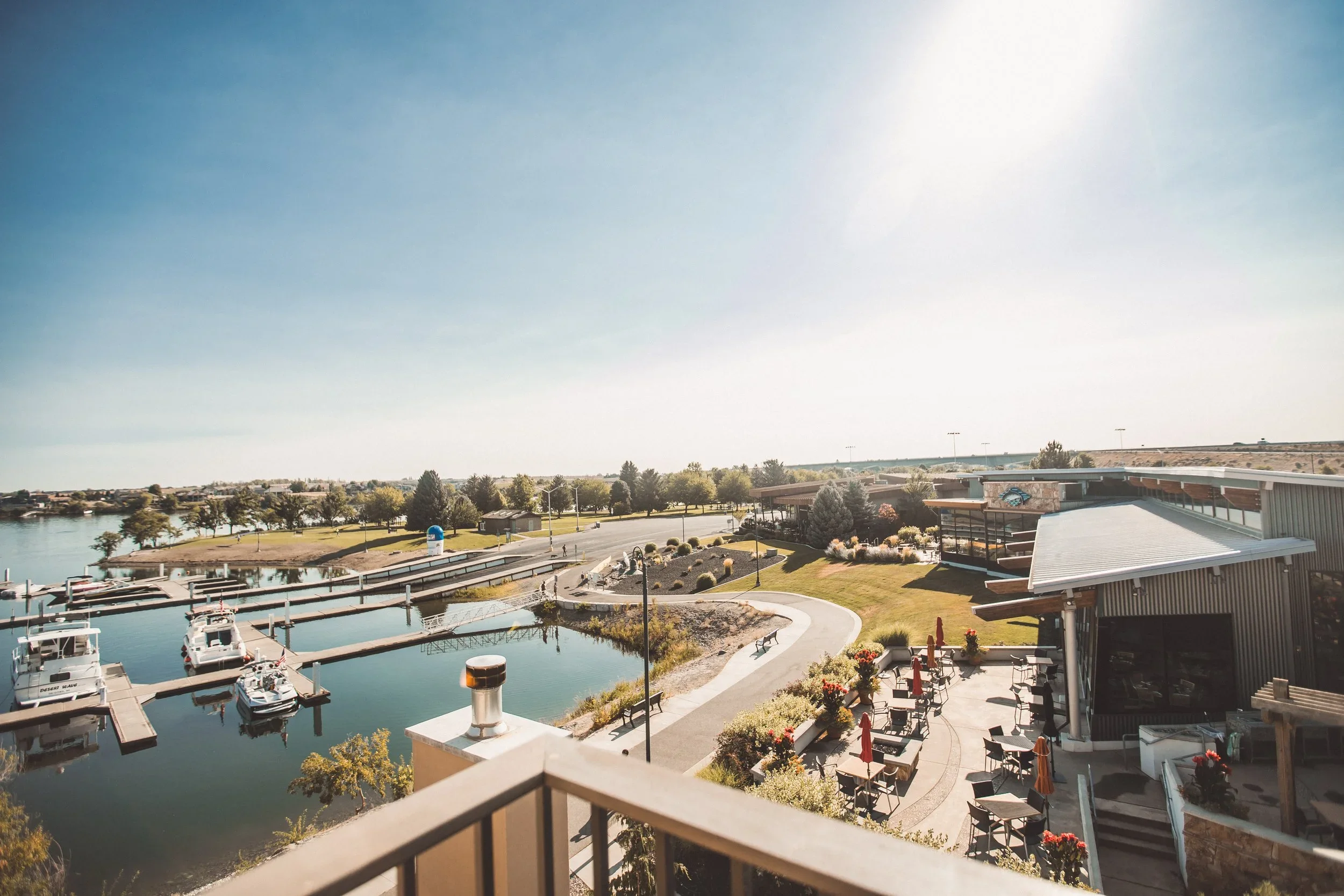 A marina with boats docked in calm water, surrounding greenery, and buildings in the background, under a clear blue sky with bright sunlight.