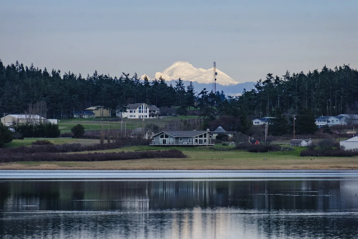 A lake with reflections in the foreground, houses and trees on the shore, a snow-capped mountain in the background, and a cloudy sky.