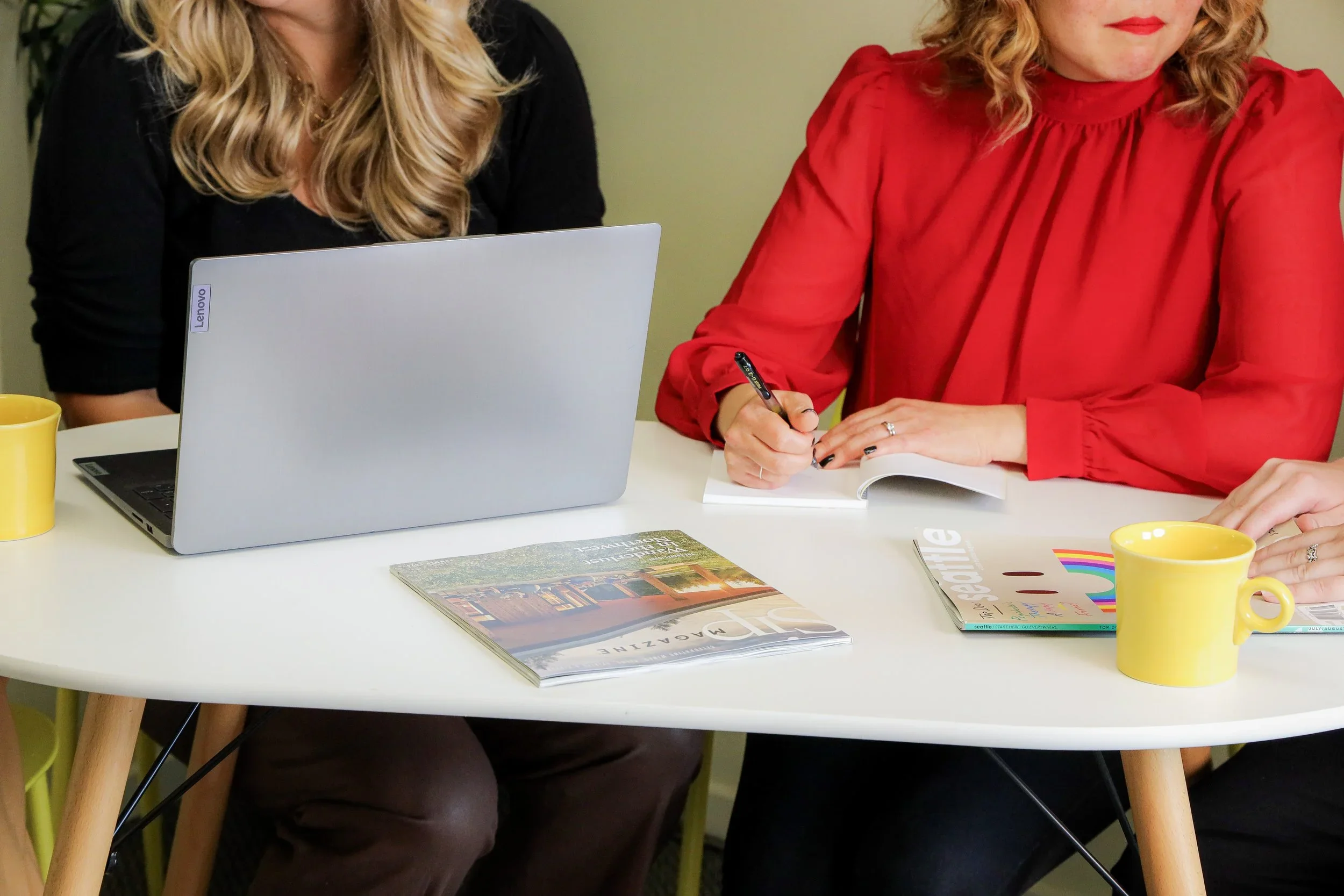 Two women sitting at a white table, one using a laptop and the other writing on a notepad. There are yellow mugs and magazines on the table, with a green wall in the background.