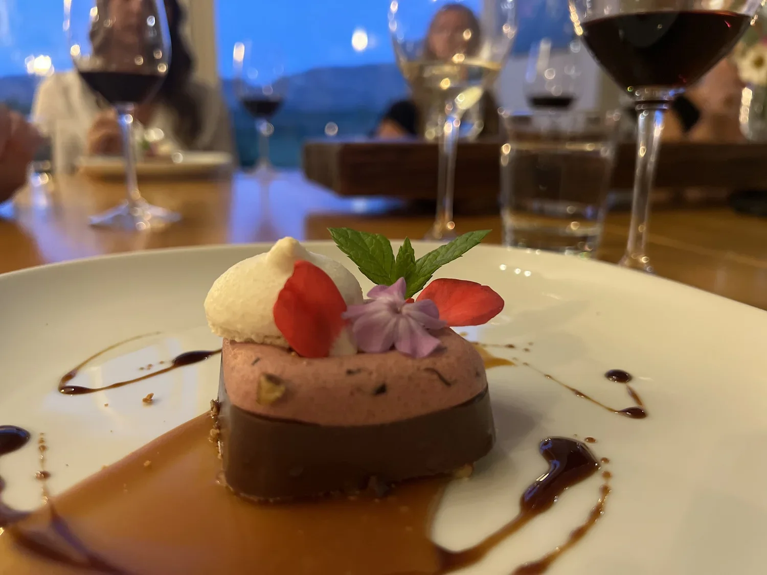 A plated dessert garnished with a mint leaf, a purple flower, strawberry slices, and a dollop of cream. The plate has decorative chocolate and caramel drizzle, with a caramel sauce underneath the dessert.