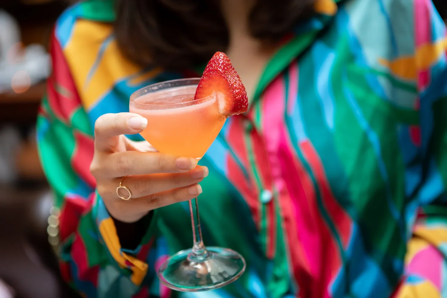 Person holding a pink cocktail with a strawberry garnish in a stemmed glass, wearing a colorful striped shirt.