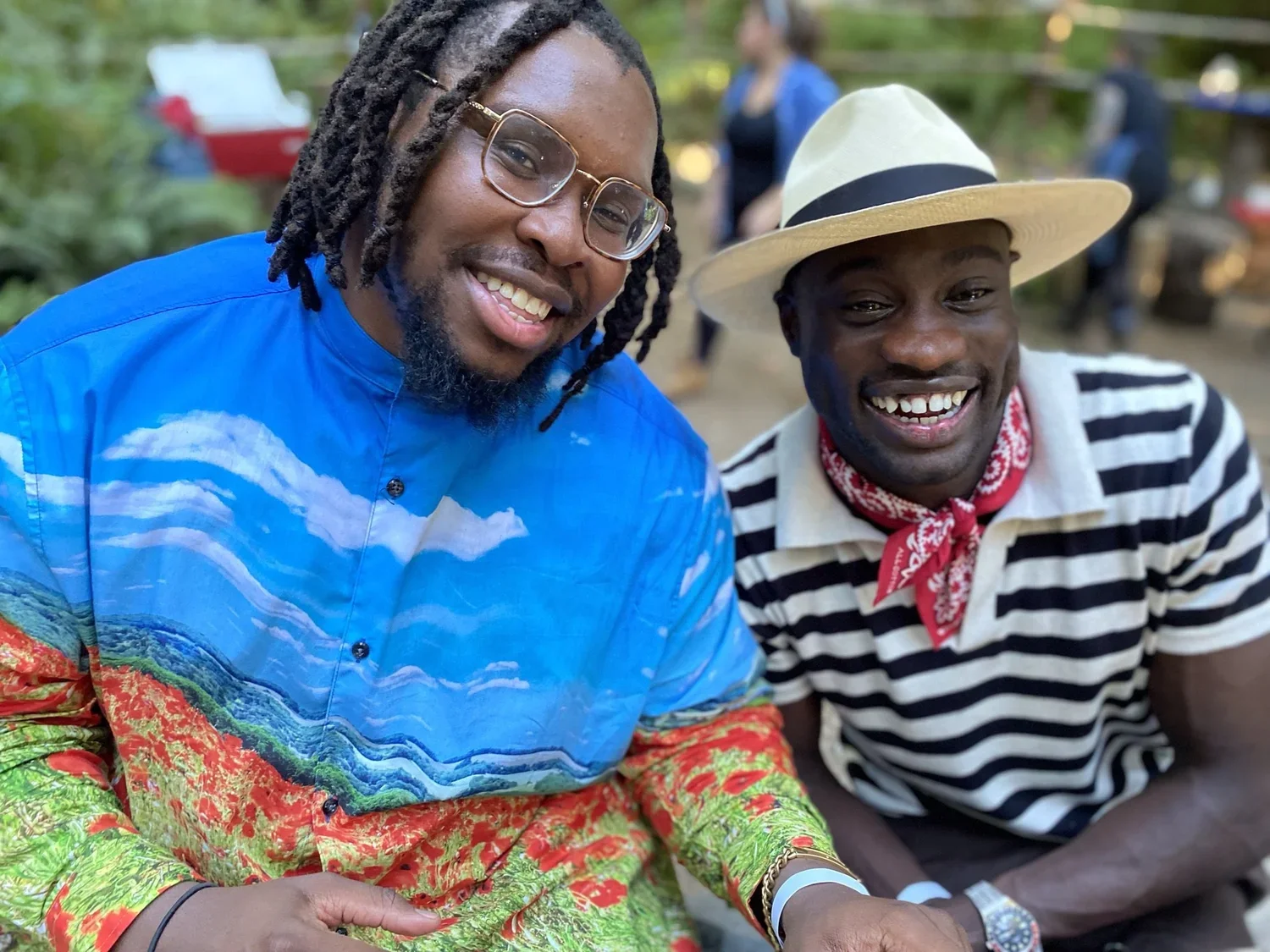 Two smiling men sitting outdoors, one wearing glasses and a colorful shirt, the other wearing a white hat, striped shirt, and red bandana.