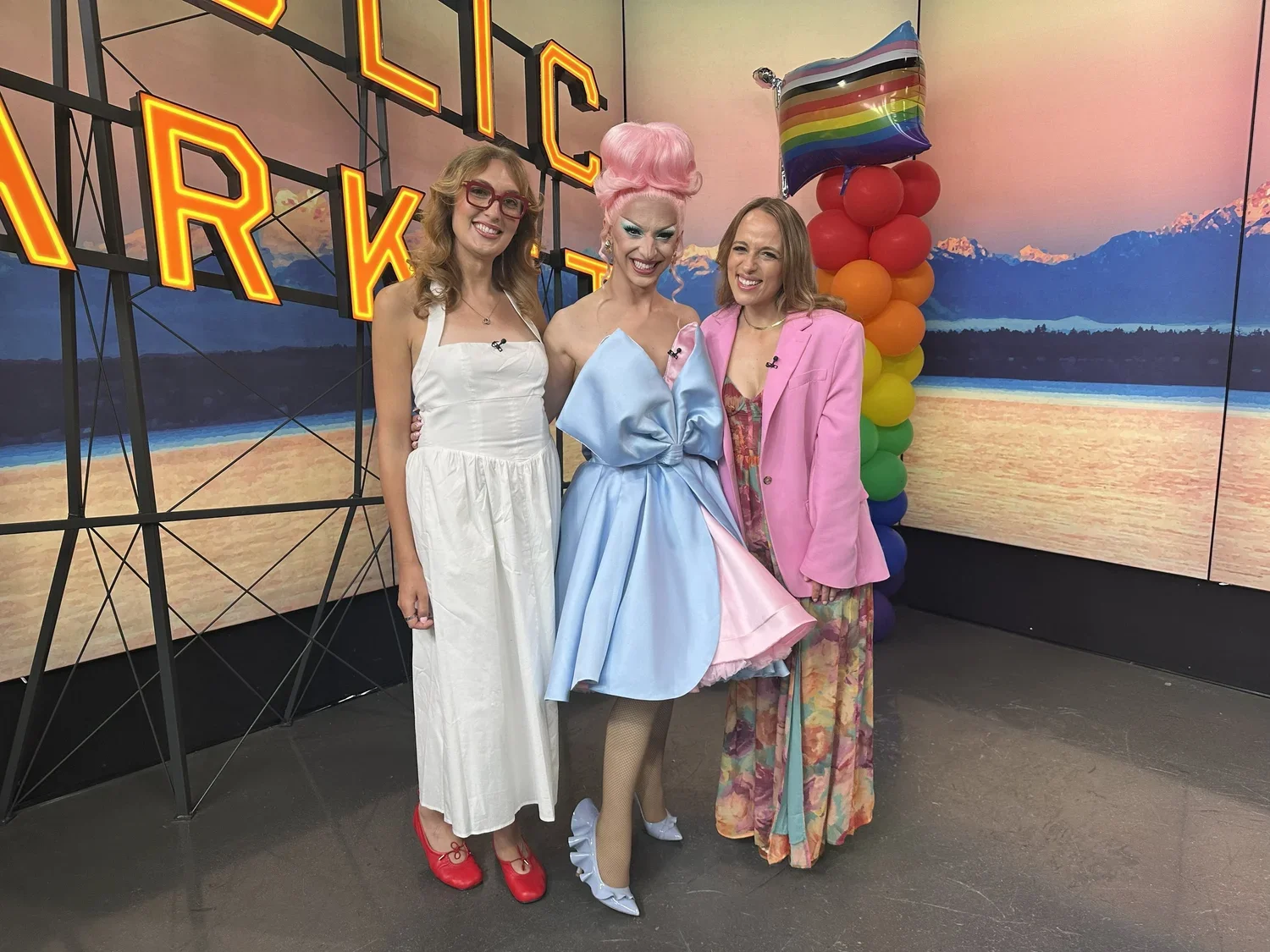 Three women standing together in front of a colorful backdrop and rainbow balloons. The woman in the middle is dressed in a big, pastel blue dress with pink hair styled in an elaborate updo. The woman on the left is wearing a white dress with red shoes and glasses. The woman on the right is wearing a pink blazer over a floral dress.