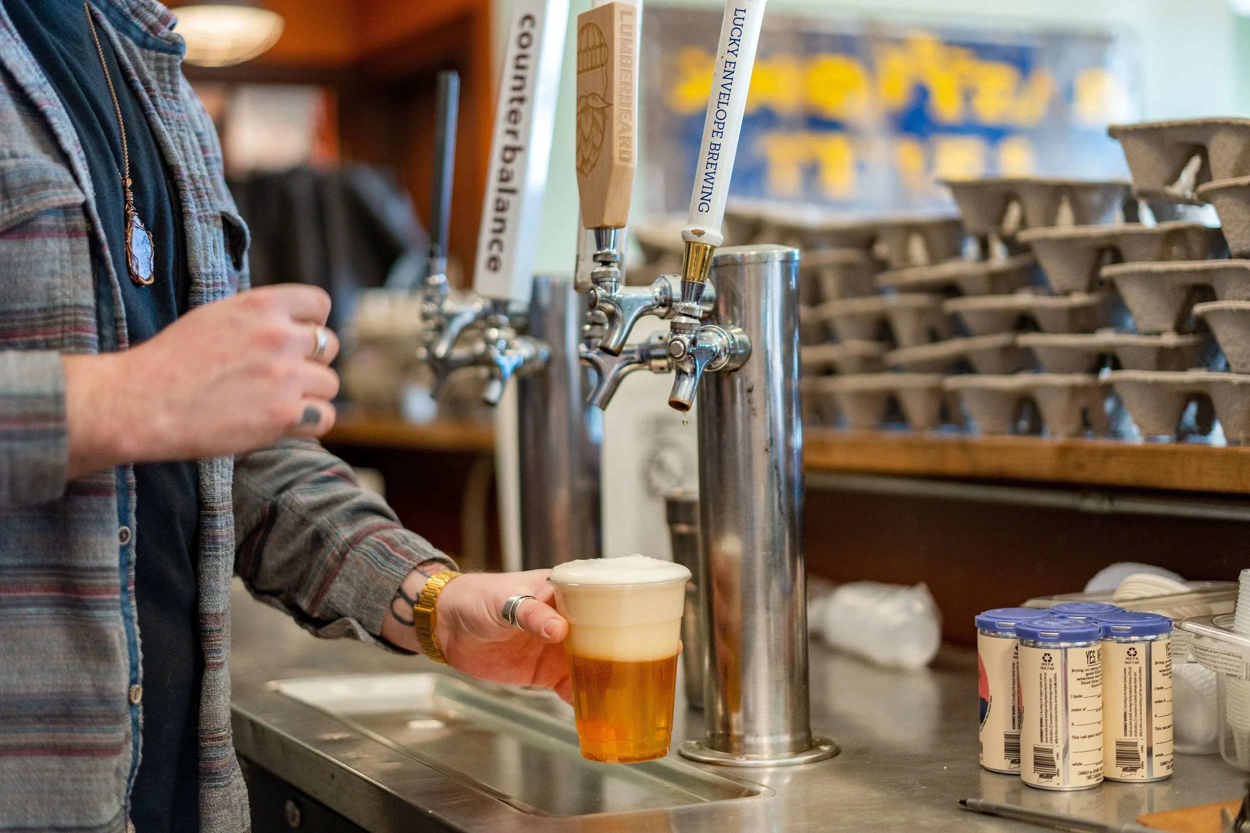 Person pouring beer from a tap into a clear plastic cup, with some bottles of yogurt on the counter nearby.