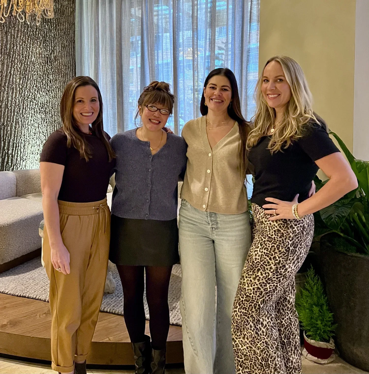Four women smiling and standing together indoors, with a tree-themed textured wall, large windows with light curtains, and plants in the background.