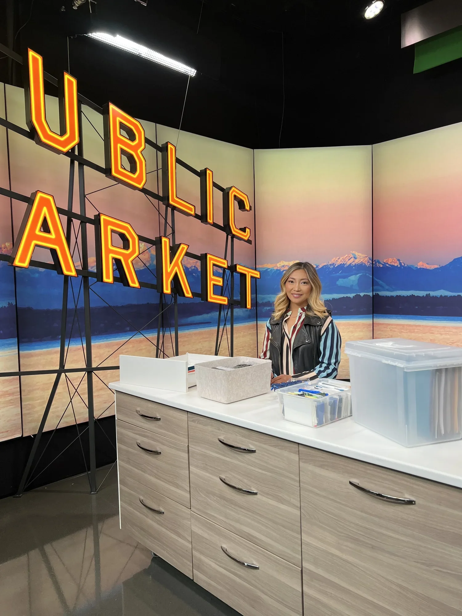 Indoor scene at a mini urban marketplace with a large neon sign reading 'URBIC MARKET' on a backdrop of mountains and sky mural. A woman is standing behind a counter with various containers and supplies, smiling at the camera.