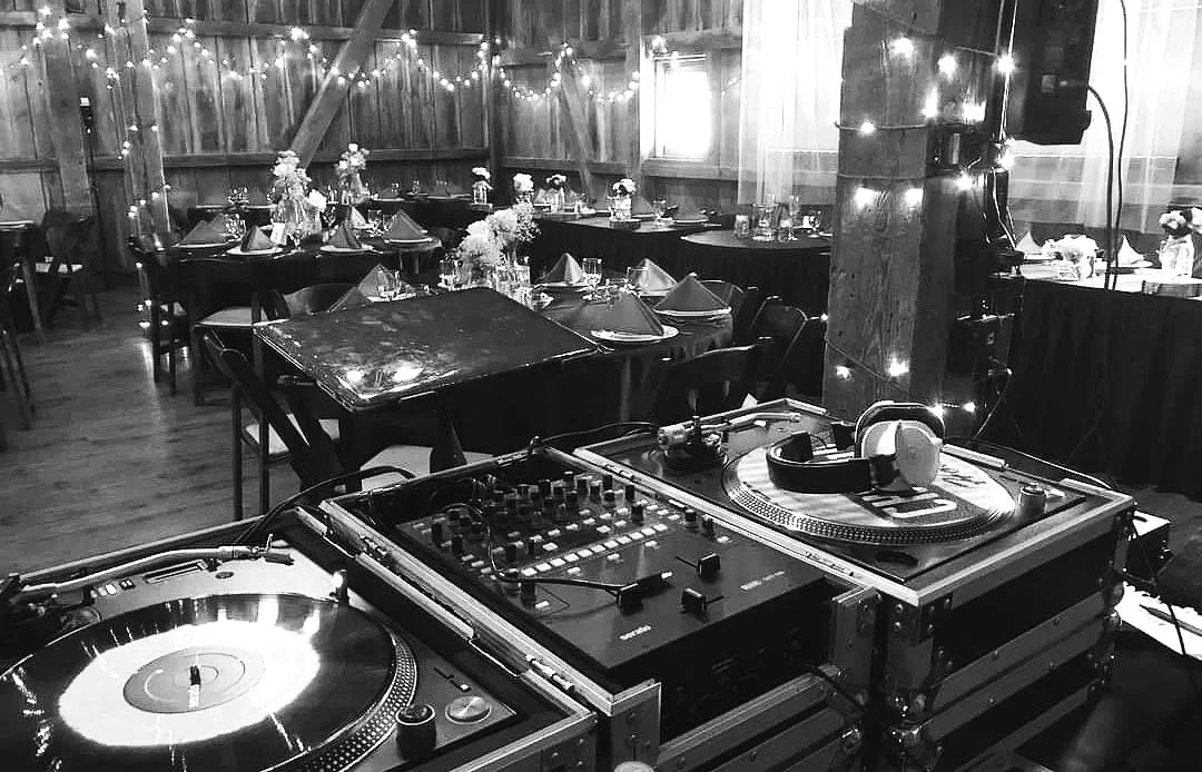 A DJ's turntables and mixing equipment set up inside a rustic barn decorated with string lights, with tables prepared for a celebration or event.