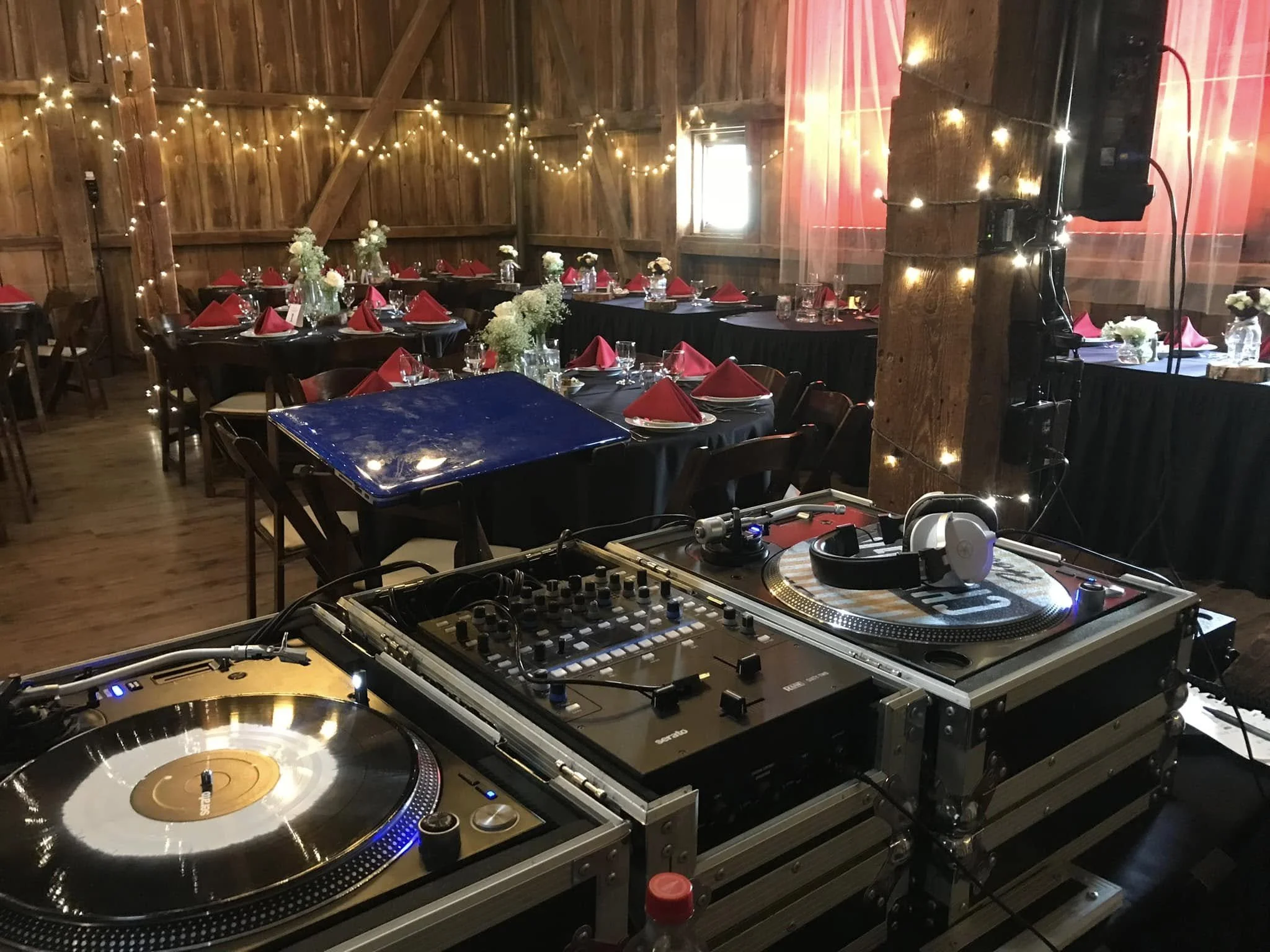 DJ equipment with turntables and mixer set up in a decorated barn for an event, with tables adorned with flowers and red napkins, and string lights hanging from wooden beams.
