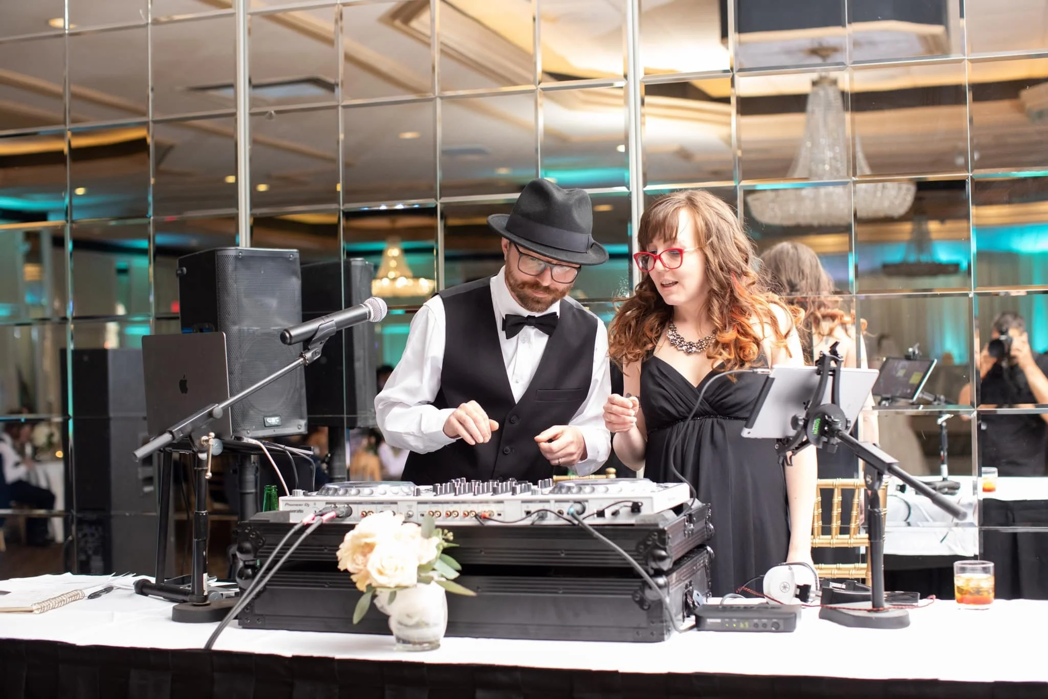 A man and woman dressed in formal attire, DJ equipment, a microphone, a tablet, and a glass of drink on a table at an event.