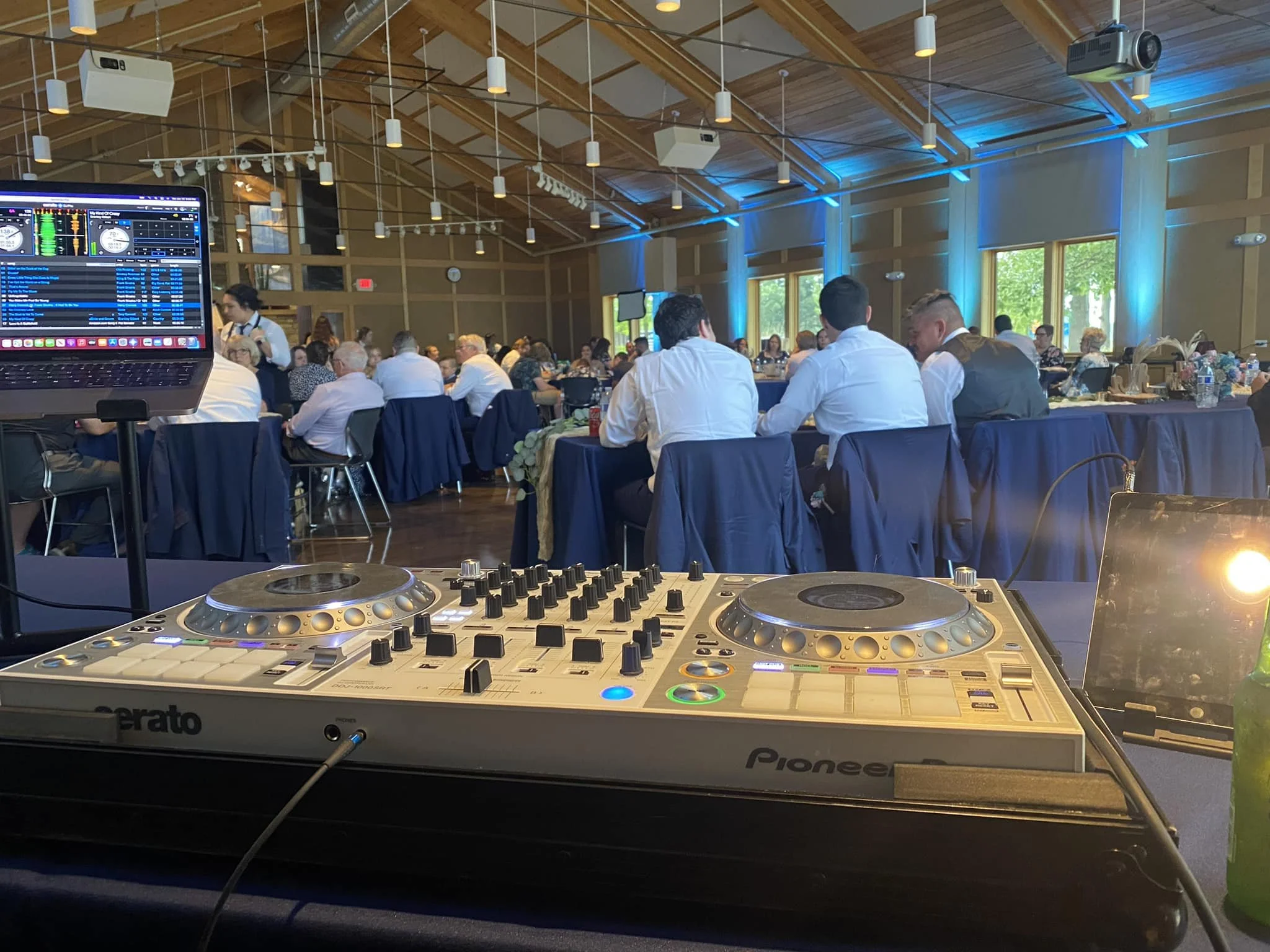 DJ equipment with a Pioneer DDJ controller and a laptop on a table at a large indoor event with people seated at round tables, decorated with tablecloths, and eating, in a wooden-ceiling venue with blue lighting and large windows.
