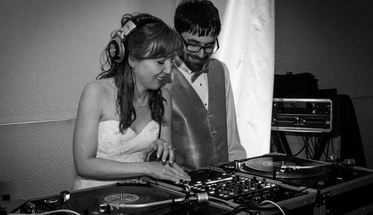 A woman and a man are DJing together at an event, with the woman wearing headphones and smiling while using turntables and a mixer, and the man standing beside her, smiling.