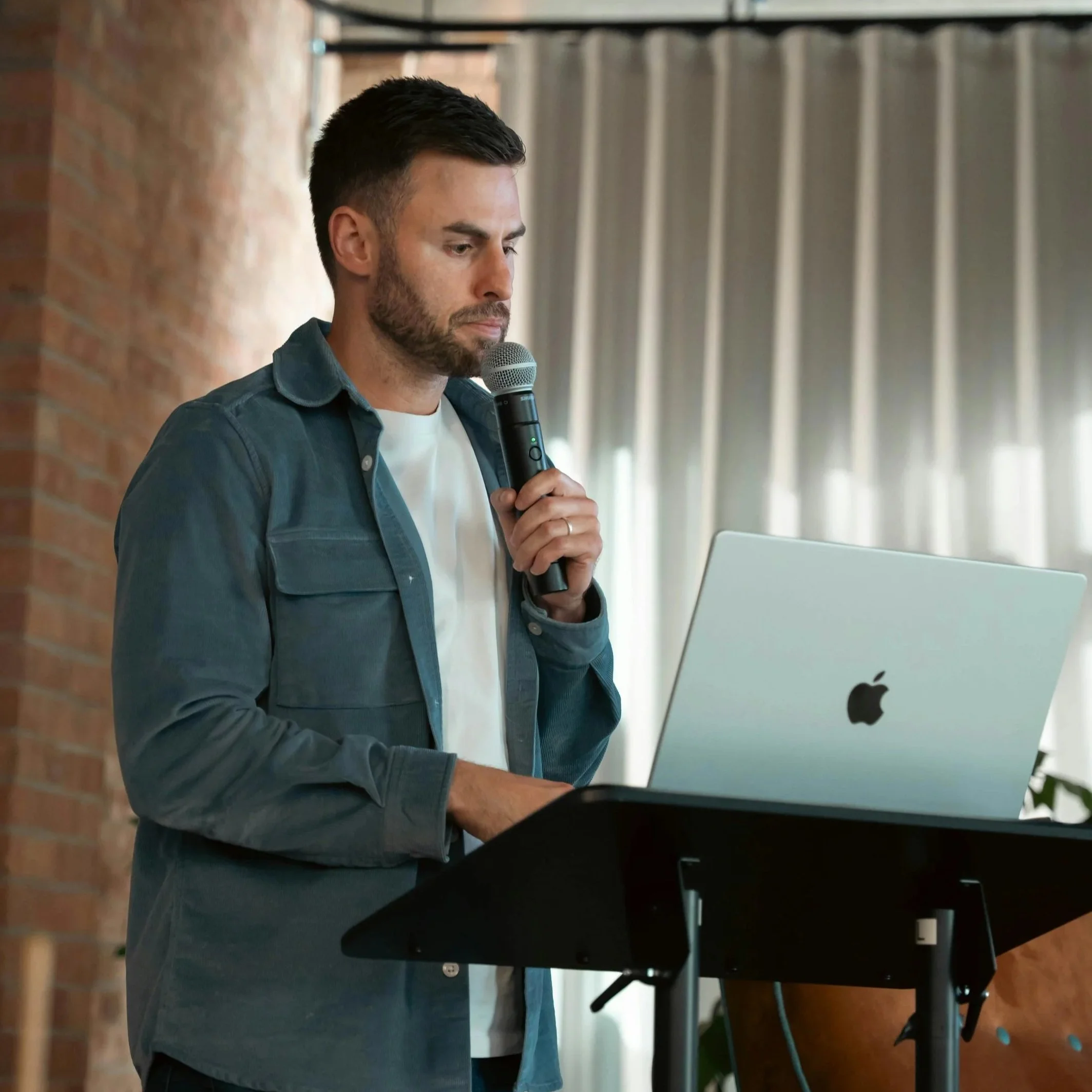 A man with dark hair and a beard is holding a microphone and looking at a laptop on a stand, in an indoor setting with brick wall and curtains in the background.