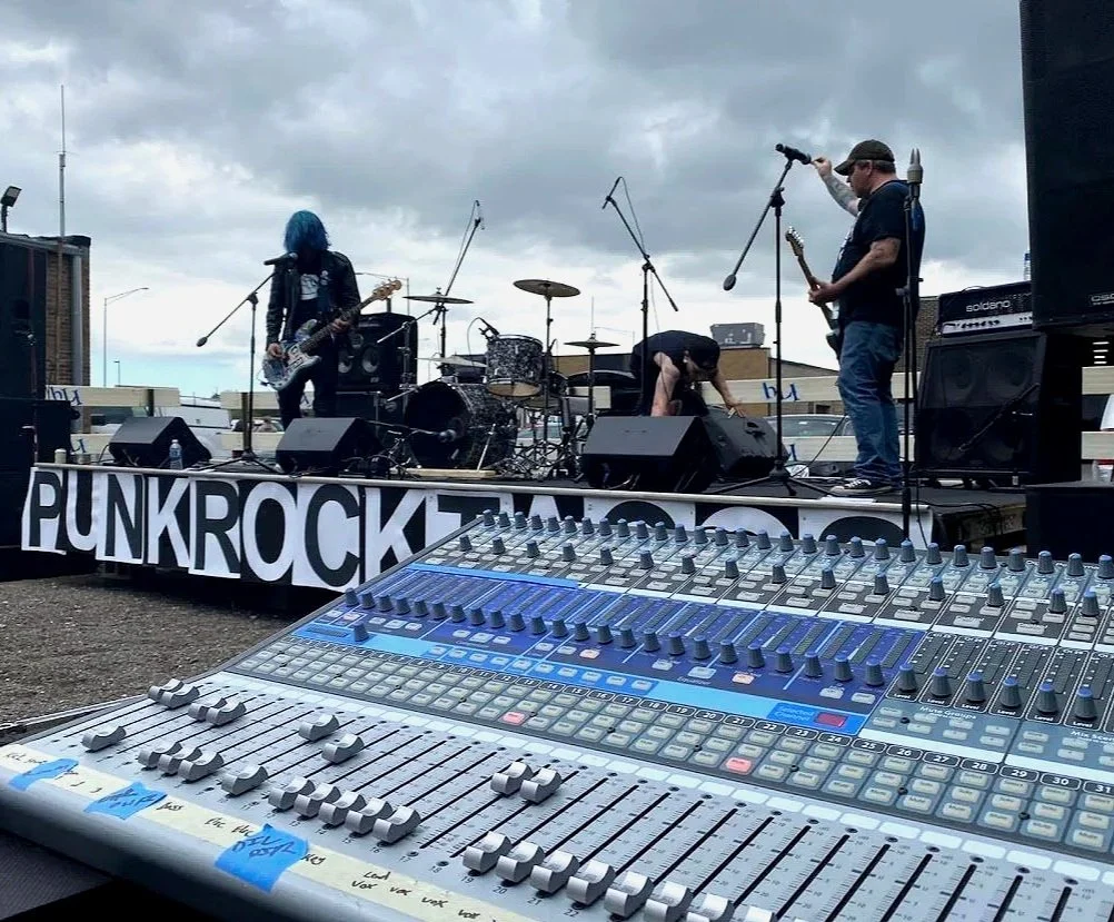 A punk rock band performing outdoors on a stage with cloudy skies; a person with blue hair playing guitar, a drummer, and two other band members; a sound mixing board in the foreground.