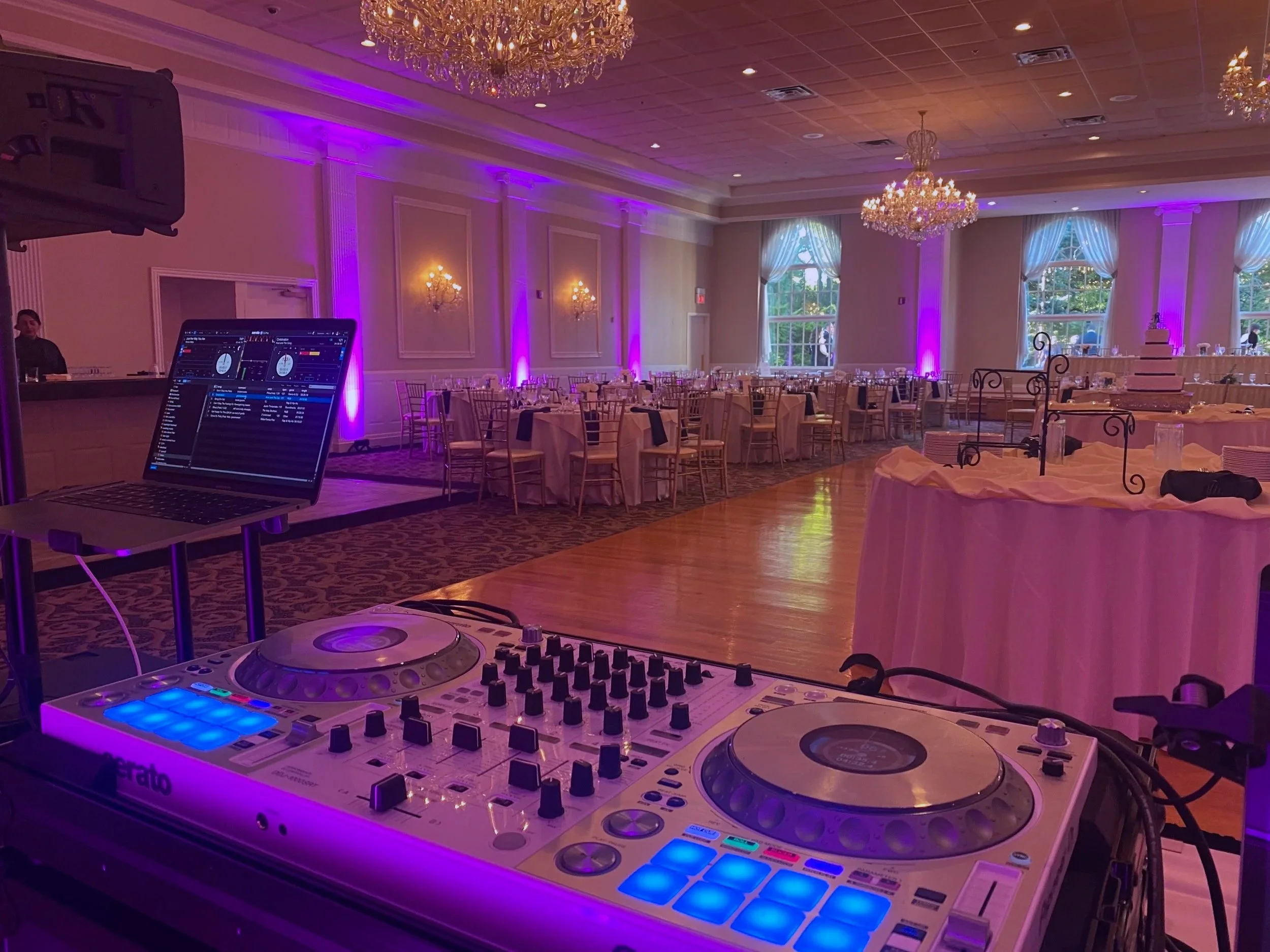 Empty banquet hall decorated with chandeliers and purple uplighting, set with round tables and chairs, with a DJ setup in the foreground including a mixer and laptop.