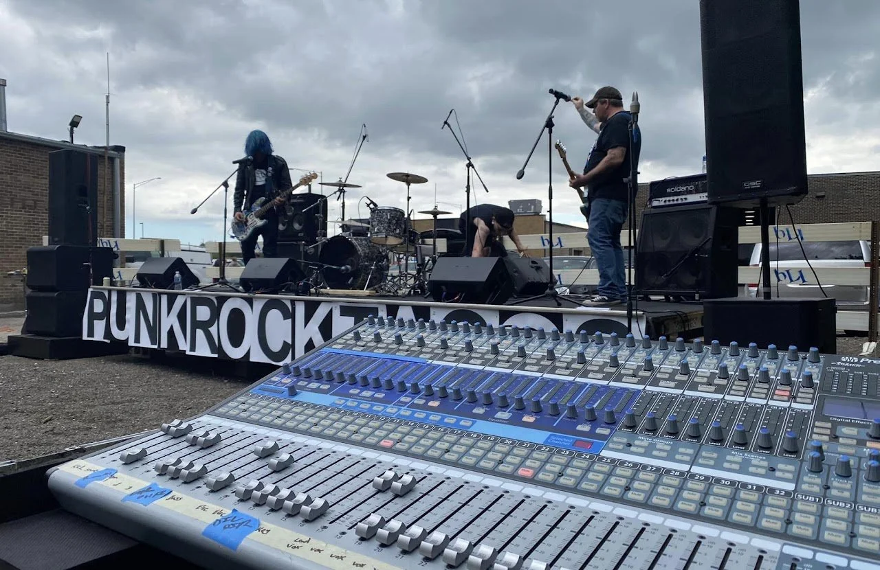 A live outdoor punk rock concert with a band performing on stage and a sound mixing console in the foreground. The stage has a banner that reads 'PUNKROCK' and features drums, guitars, and microphones. Dark clouds are overhead.