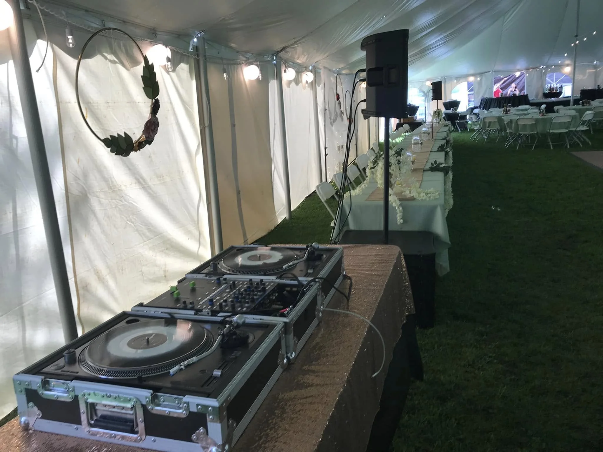 DJ equipment with turntables and mixer set up on a table inside a decorated event tent, with chairs and tables in the background.