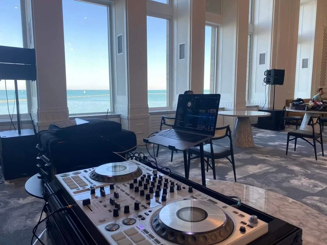 DJ equipment with turntables and a laptop on a table in a bright room with large windows showing a view of the ocean.