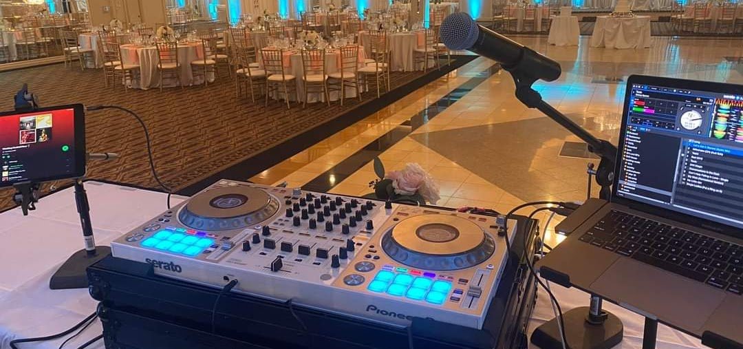 DJ equipment setup with a laptop, a microphone, and a small screen, set up on a table in an empty banquet hall with decorated tables and chairs in the background.