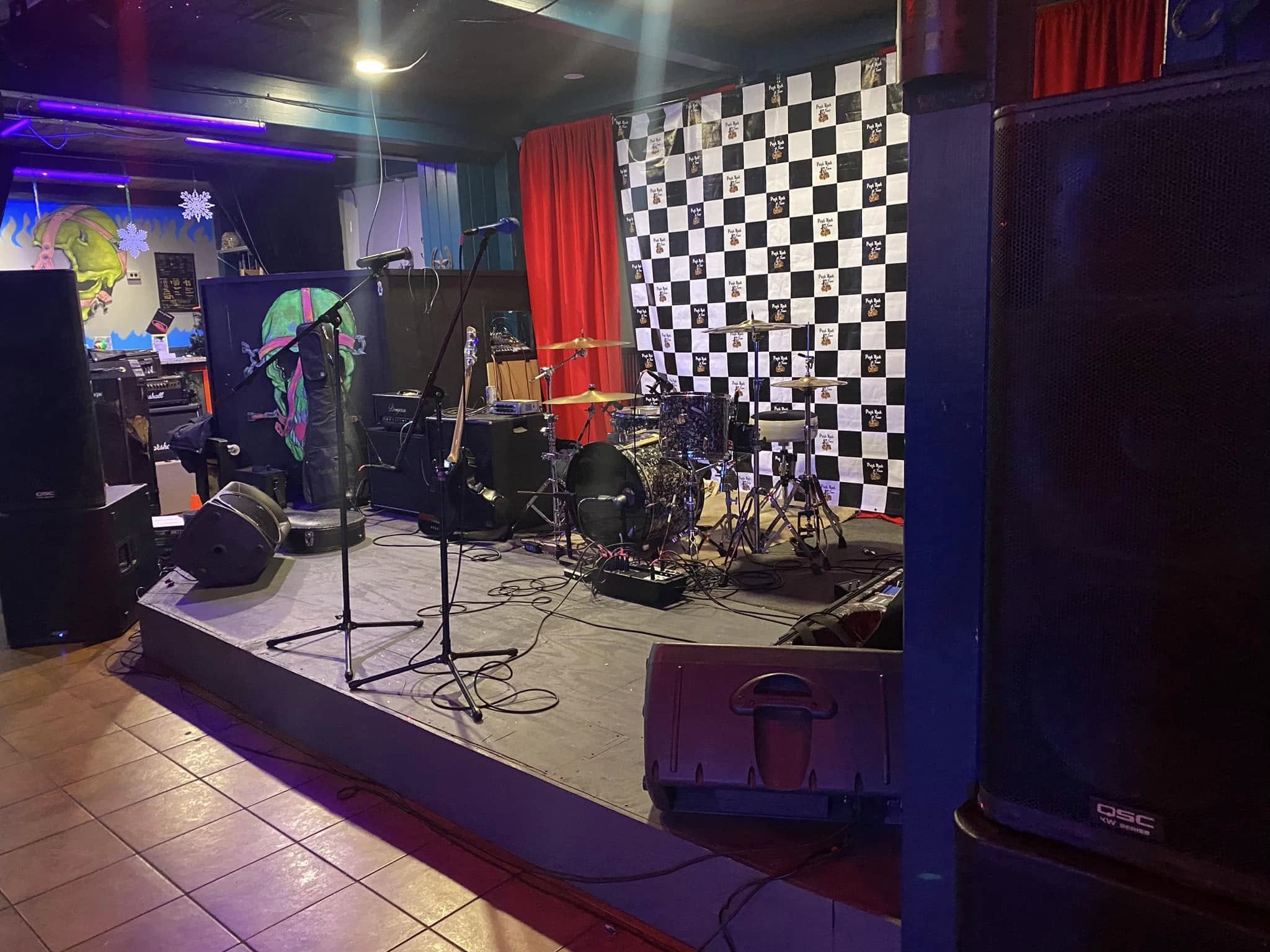 Empty music stage with drums, microphones, and speakers in a colorful club with red curtains and a black-and-white checkered backdrop.