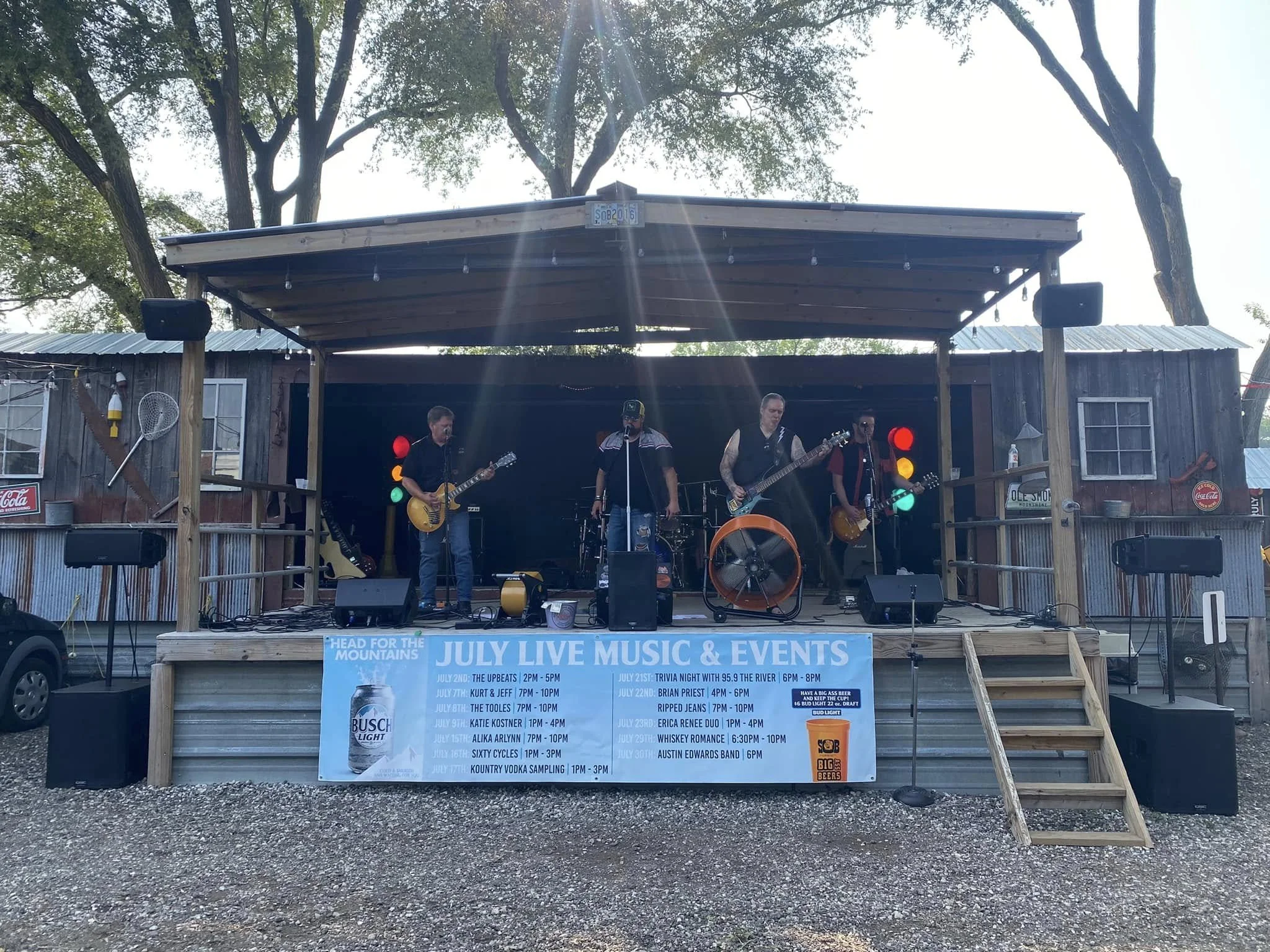 A live outdoor band performing on a stage with a banner listing July events, with trees in the background and some gravel on the ground.