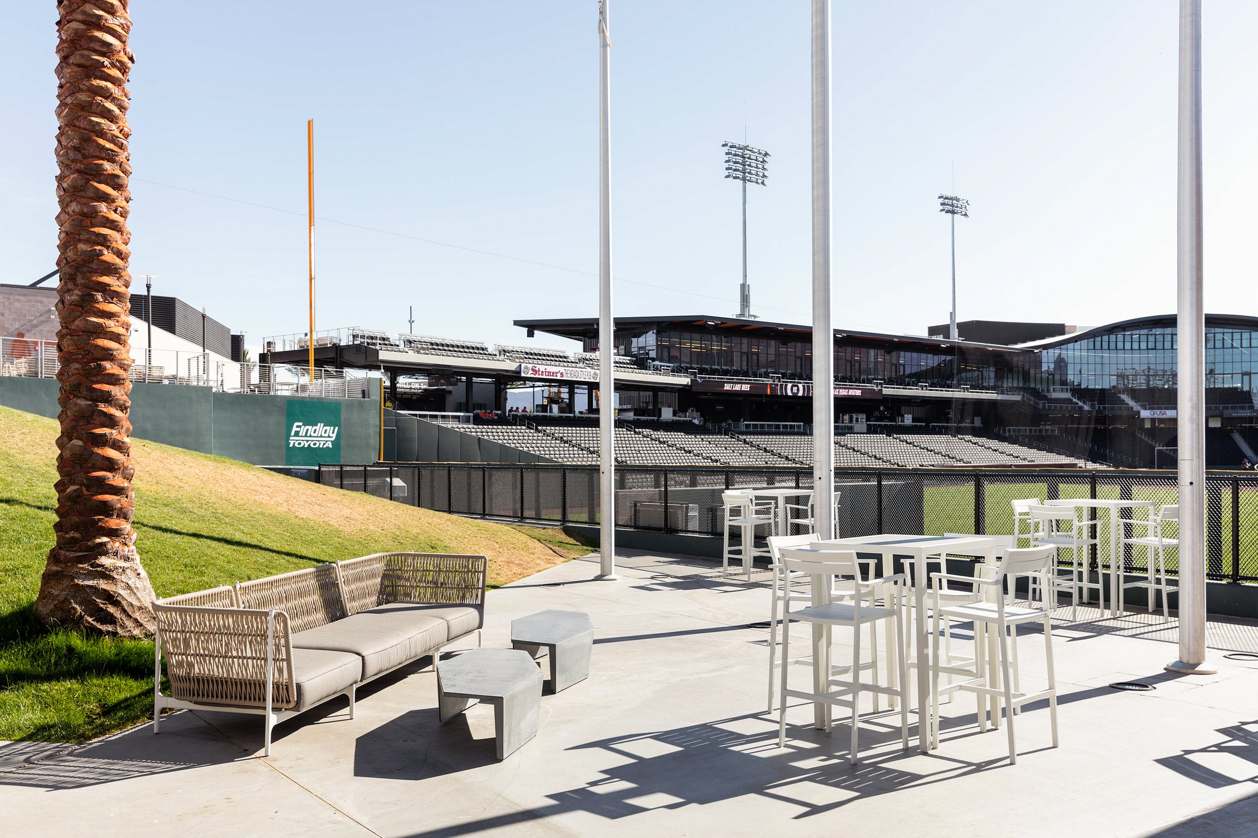 Outdoor seating area at a sports stadium with white lawn chairs and a beige couch, a palm tree on the left, and a fenced off field with stadium stands and tall light poles in the background.