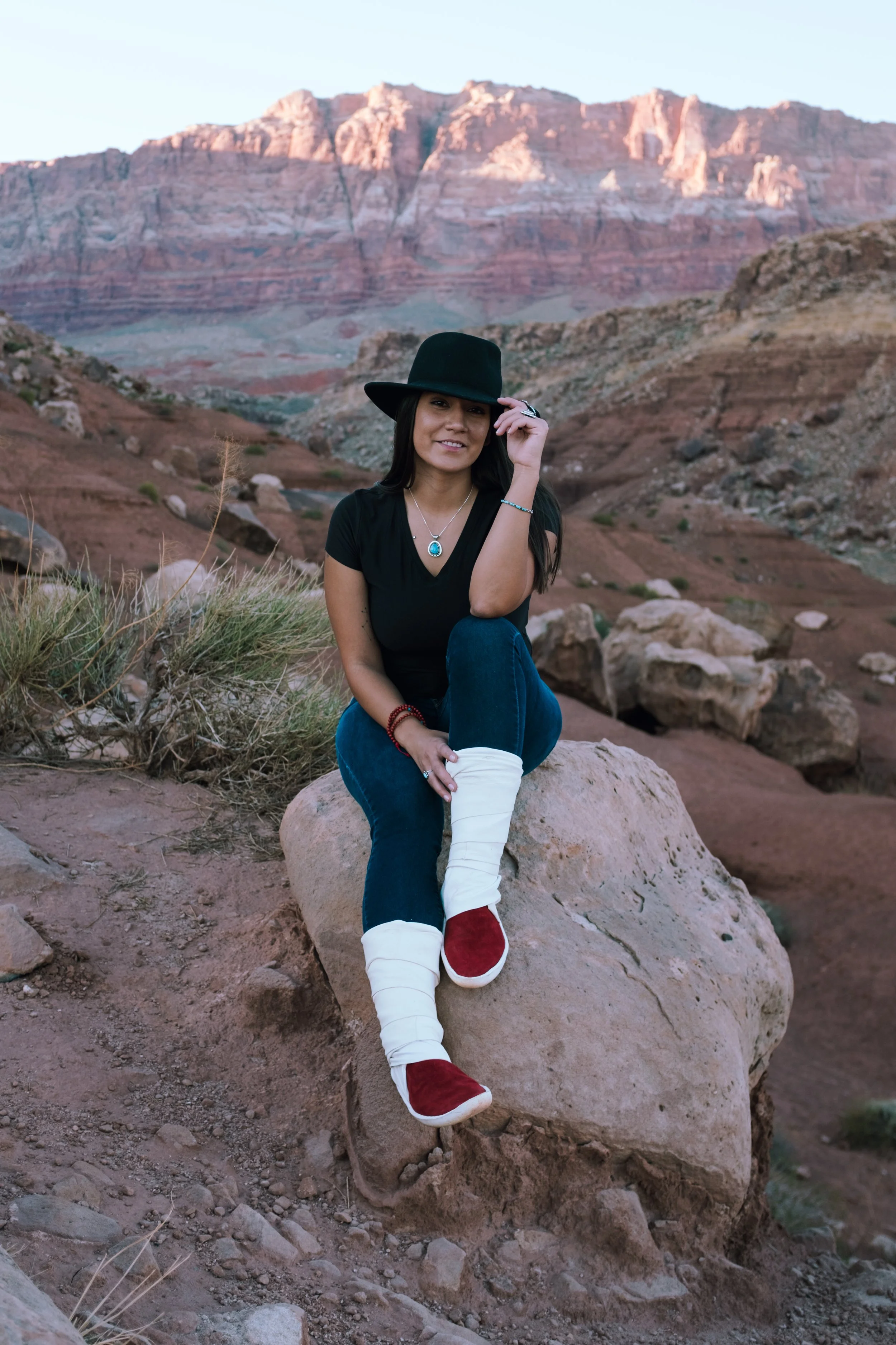 A woman sitting on a rock in a desert landscape with red rock formations in the background, wearing a black hat, black shirt, blue jeans, and red and white boots.
