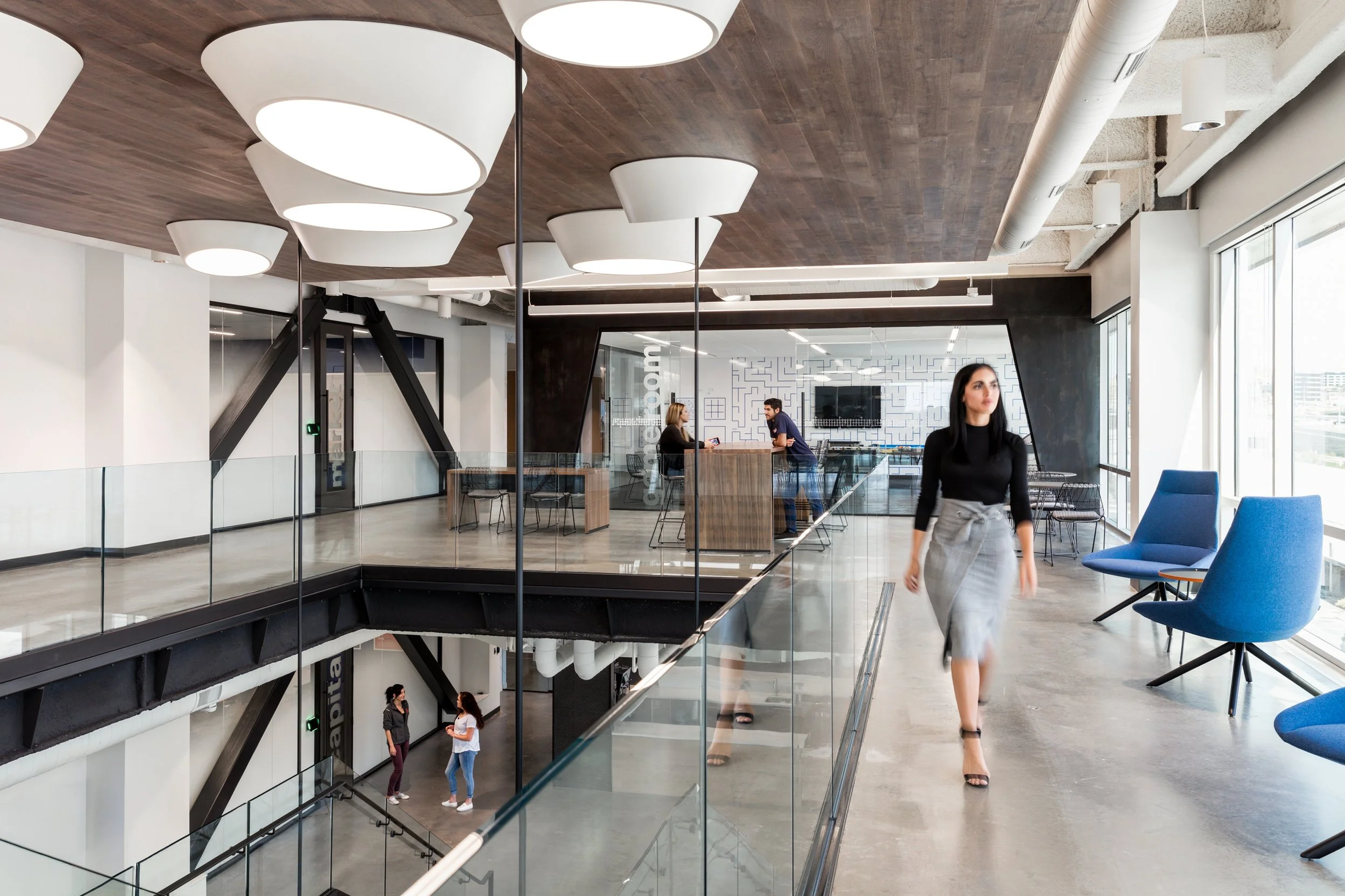 Interior of a modern office building featuring a glass-walled meeting area with four people inside, a woman walking past in the foreground, contemporary furniture, large windows, and ceiling lights.