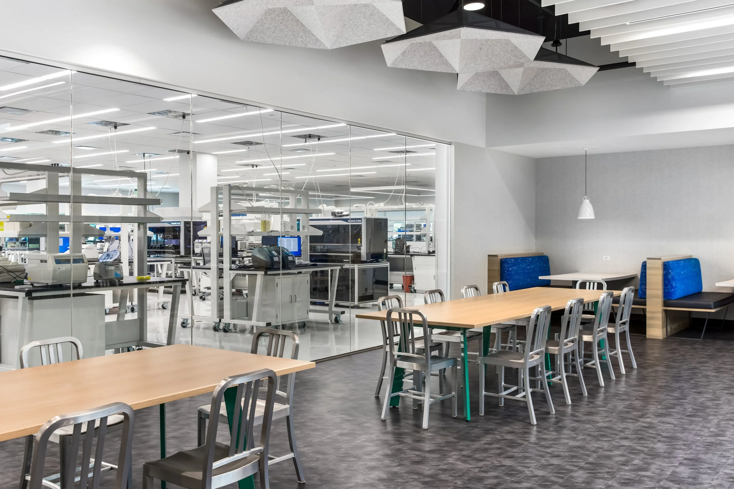 A modern, clean laboratory with glass walls, white counters, various scientific equipment, and a seating area with tables and chairs.