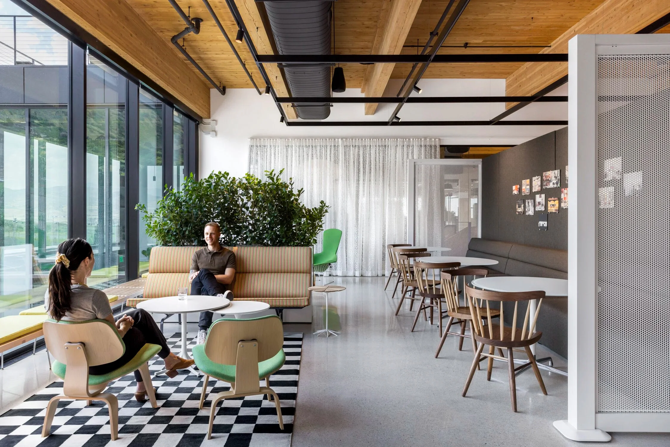 Modern coworking space with large windows, wooden ceiling, and a mix of seating arrangements. Two women are sitting at a table and talking, while a man sits on a striped sofa. There are several chairs, tables, and a photo display on the wall.