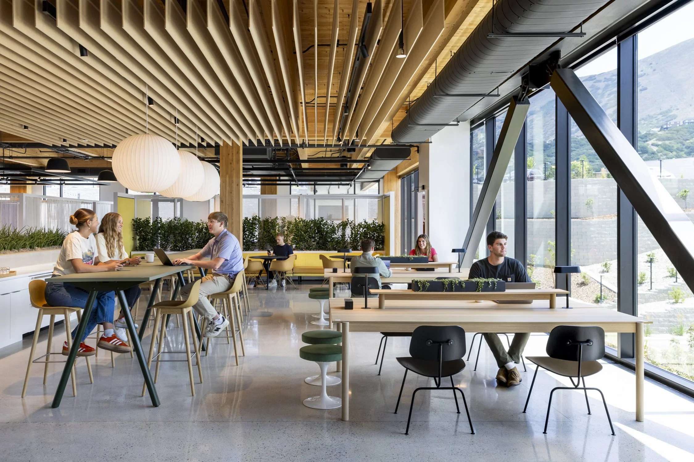 Modern coworking space with large windows, wooden ceiling with slats, hanging spherical light fixtures, and people working at tables.