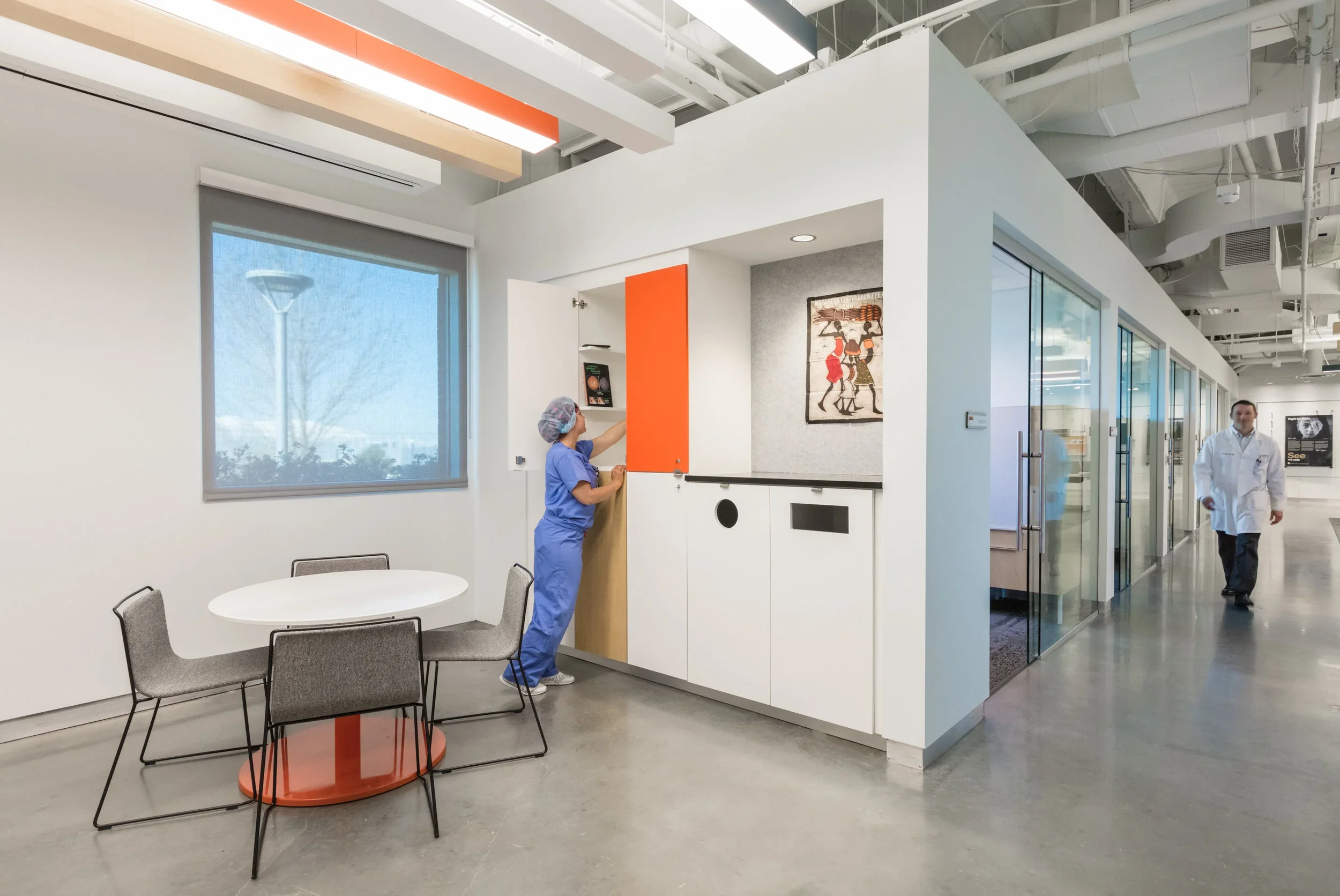 Inside a modern medical facility with white walls and large windows. A healthcare worker in scrubs and a hairnet is opening a door, and another person in a lab coat is walking in the hallway.