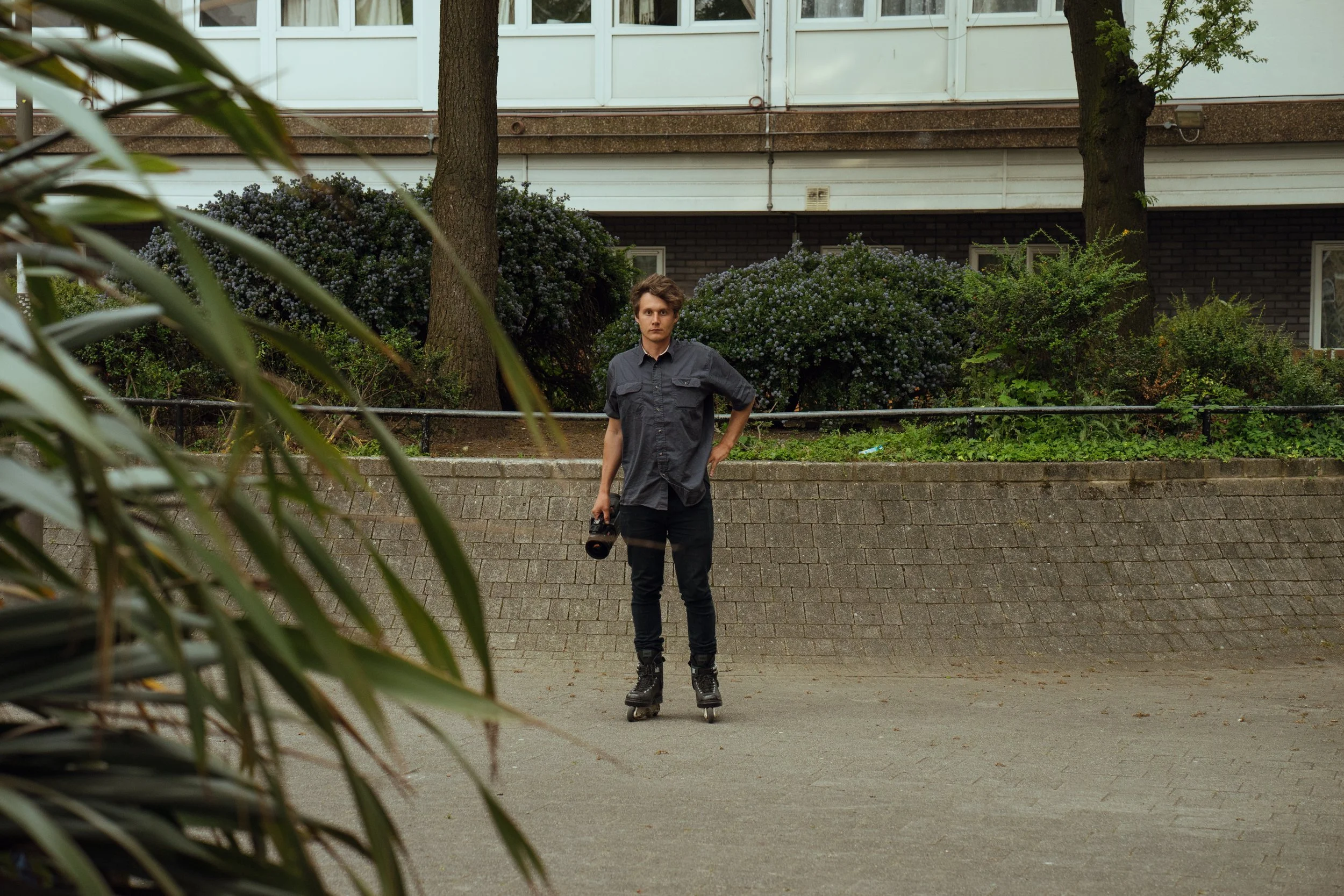 A young man standing on rollerblades in a park with trees and bushes in the background, holding a camera, wearing a gray shirt and black pants.