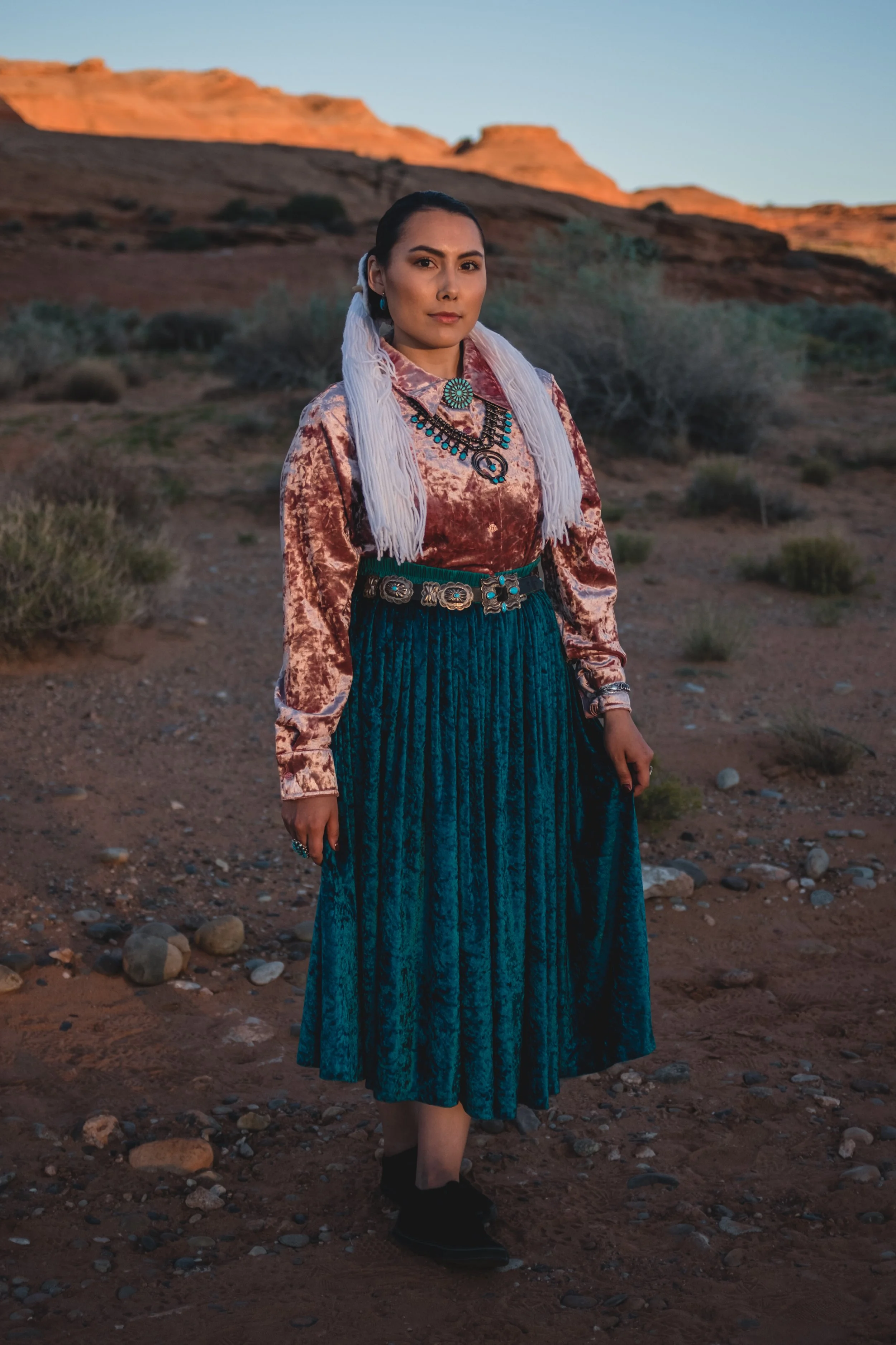 A woman dressed in traditional Native American clothing stands outdoors in a desert landscape with rock formations in the background, at sunset.