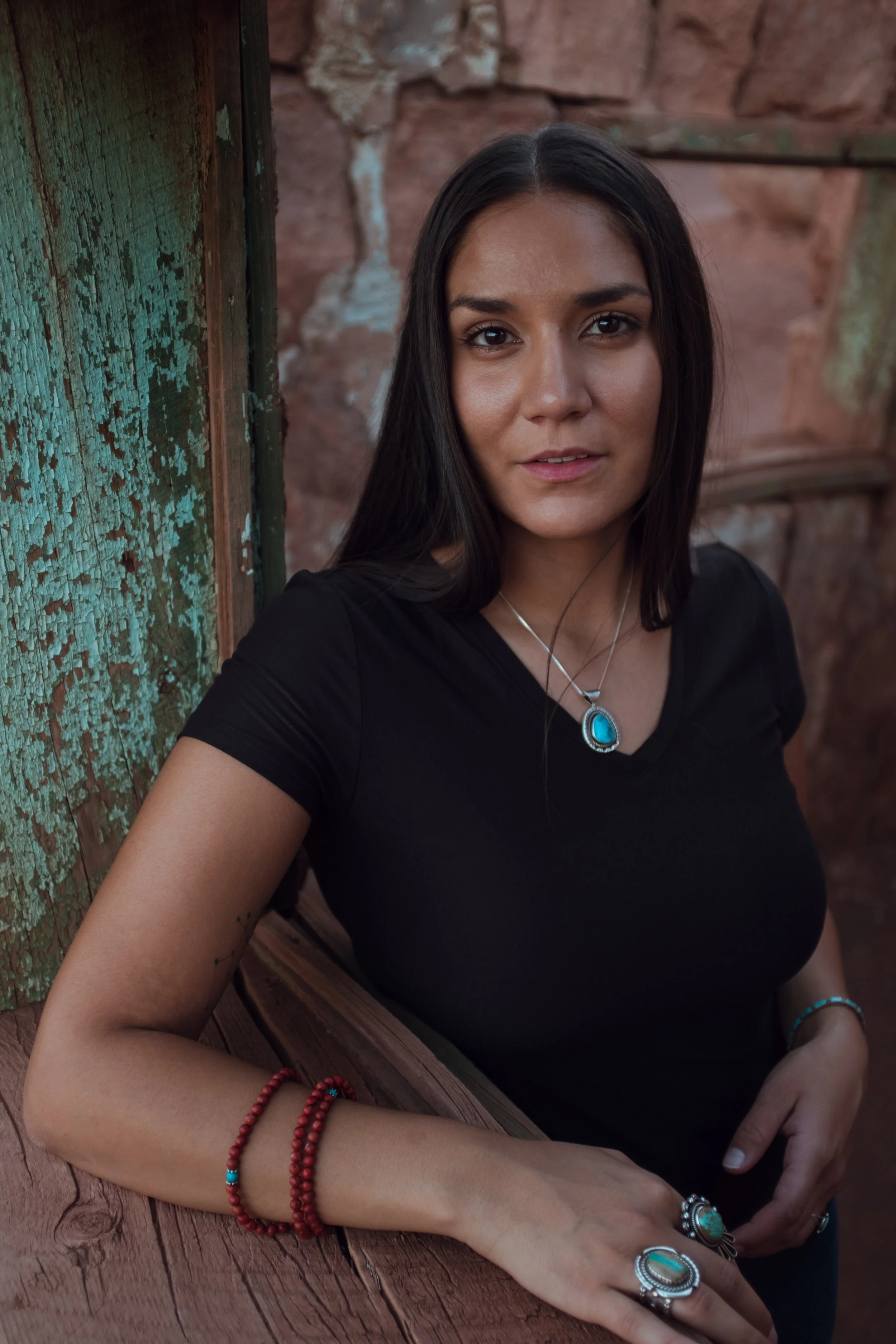 A woman with long dark hair, wearing a black shirt, turquoise jewelry, and red beaded bracelets, leaning on a wooden surface with a rustic, brick background.