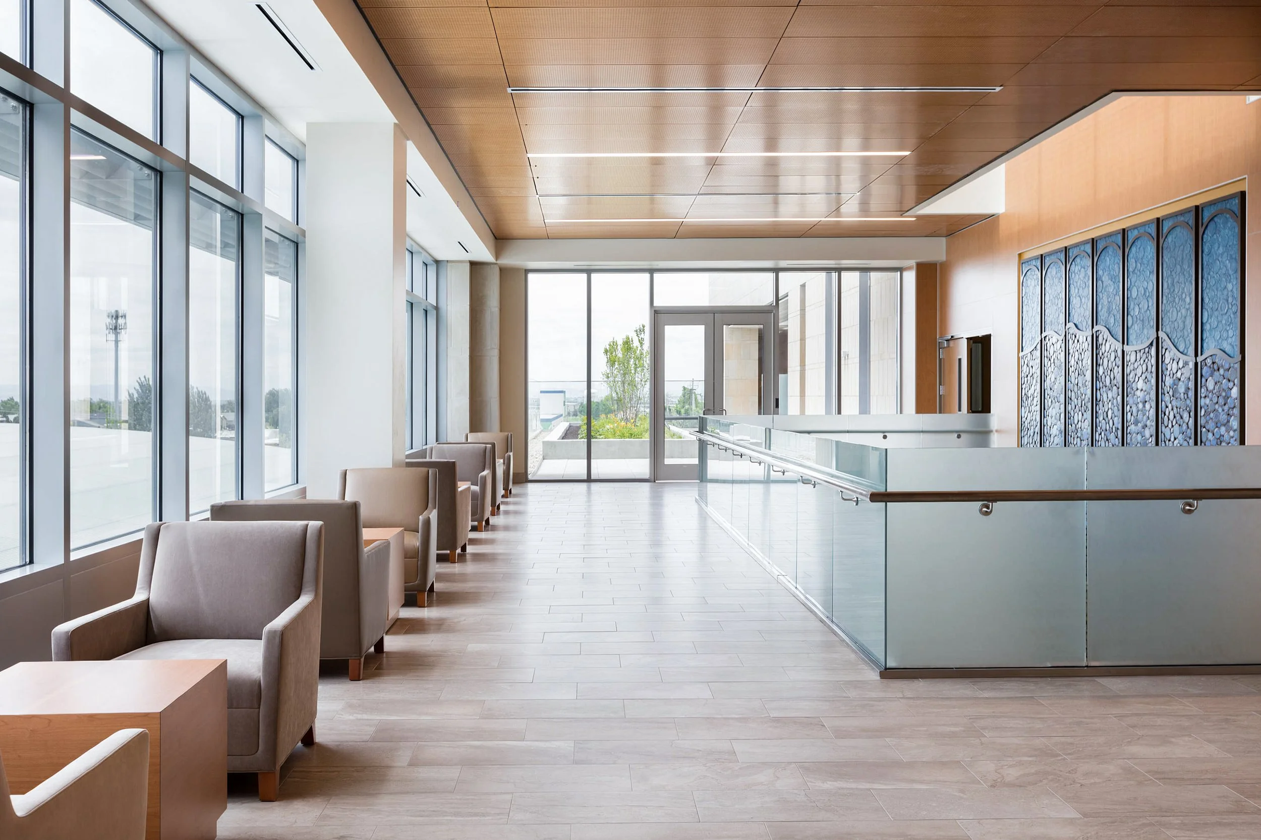 Modern lobby with large floor-to-ceiling windows, beige chairs, wooden ceiling, glass railing, and decorative blue wall panels.