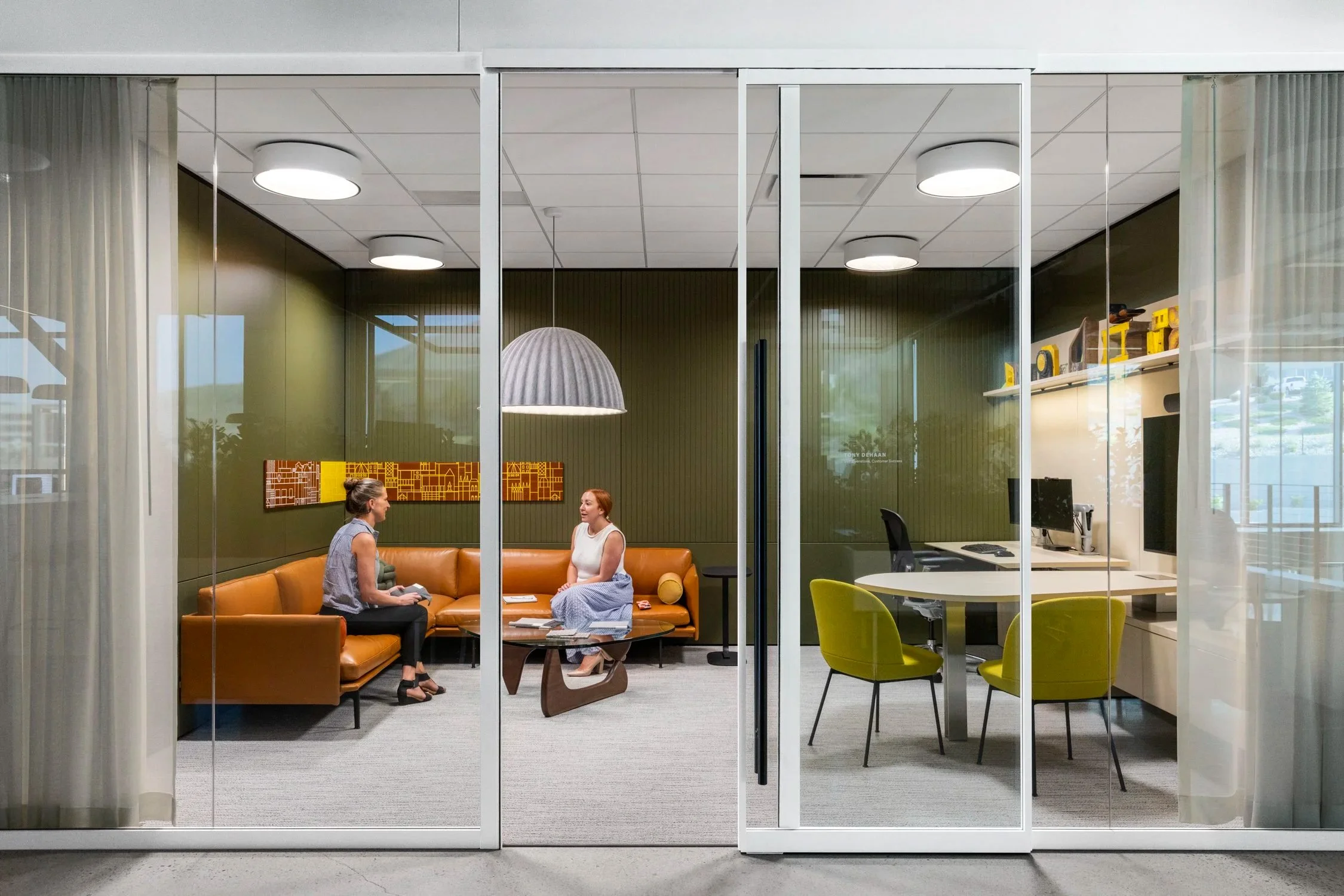 Two women having a conversation in a contemporary office lounge with glass walls, modern lighting, and colorful chairs.