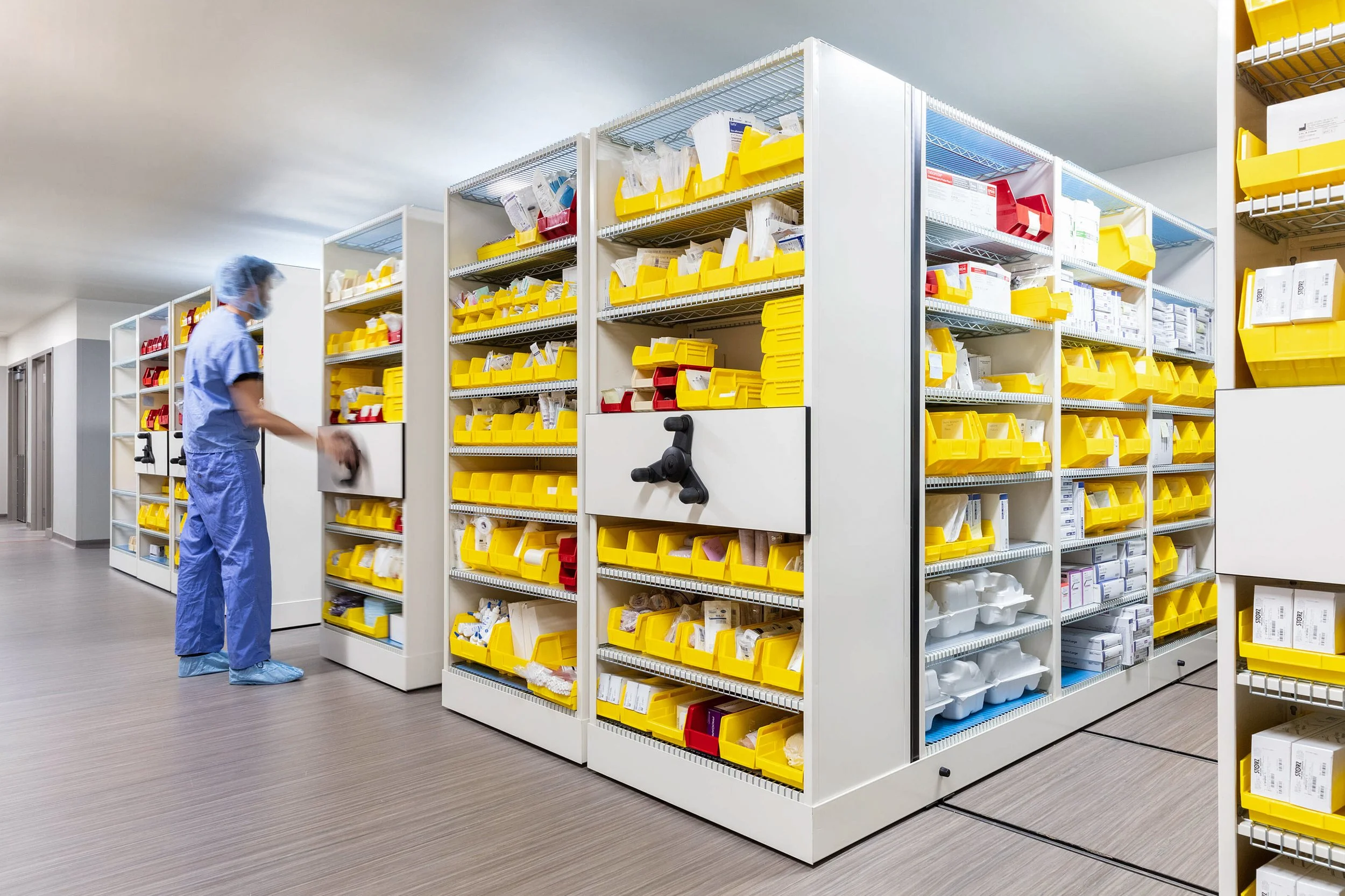 Hospital or medical supply storage area with shelves filled with yellow and red bins containing medical supplies. A worker in blue medical scrubs and a hairnet is seen organizing or retrieving supplies from the shelves.